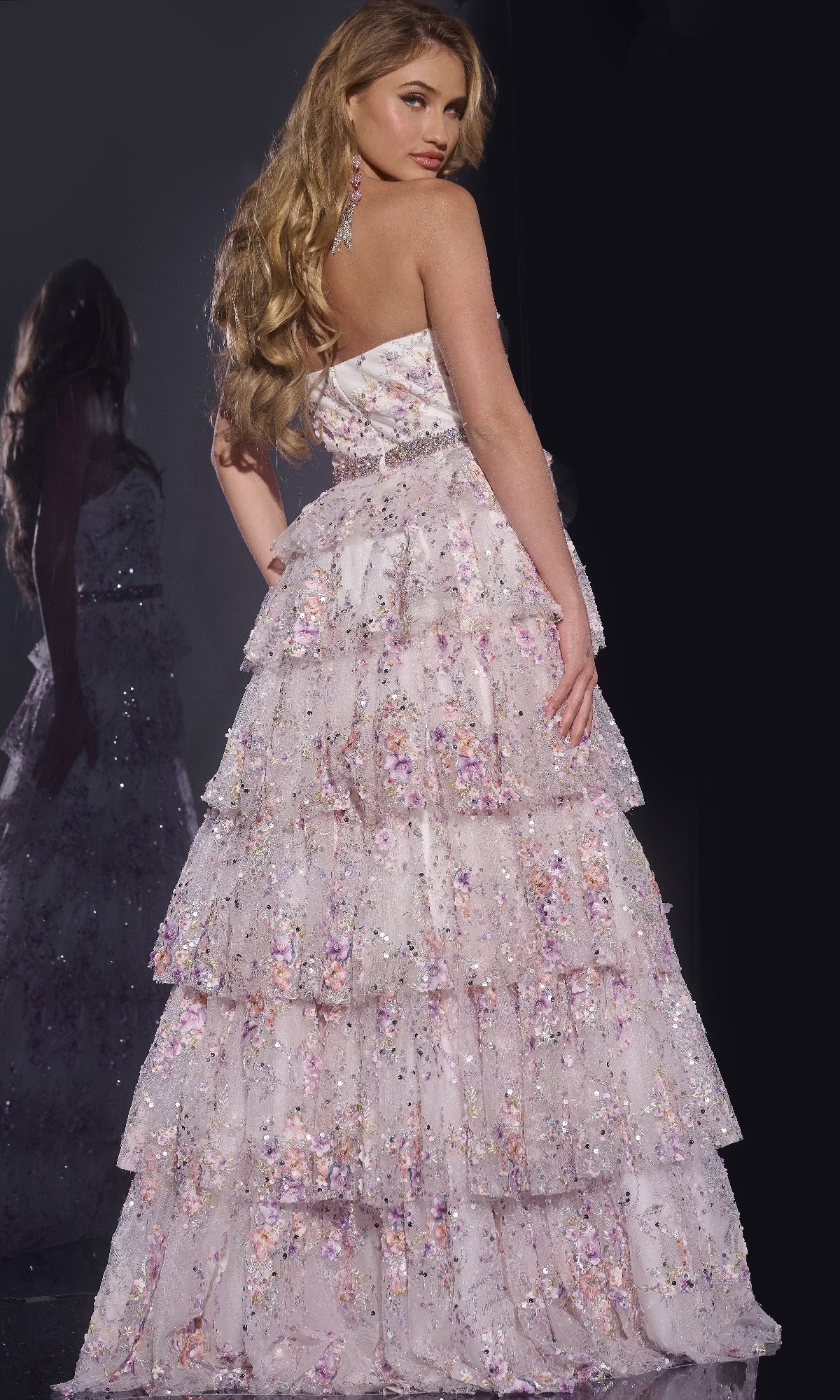 A woman with long wavy hair models the Jovani 42190, a strapless floral lace prom dress with a beaded waist and tiered, floor-length sequin skirt, posing against a dark background.