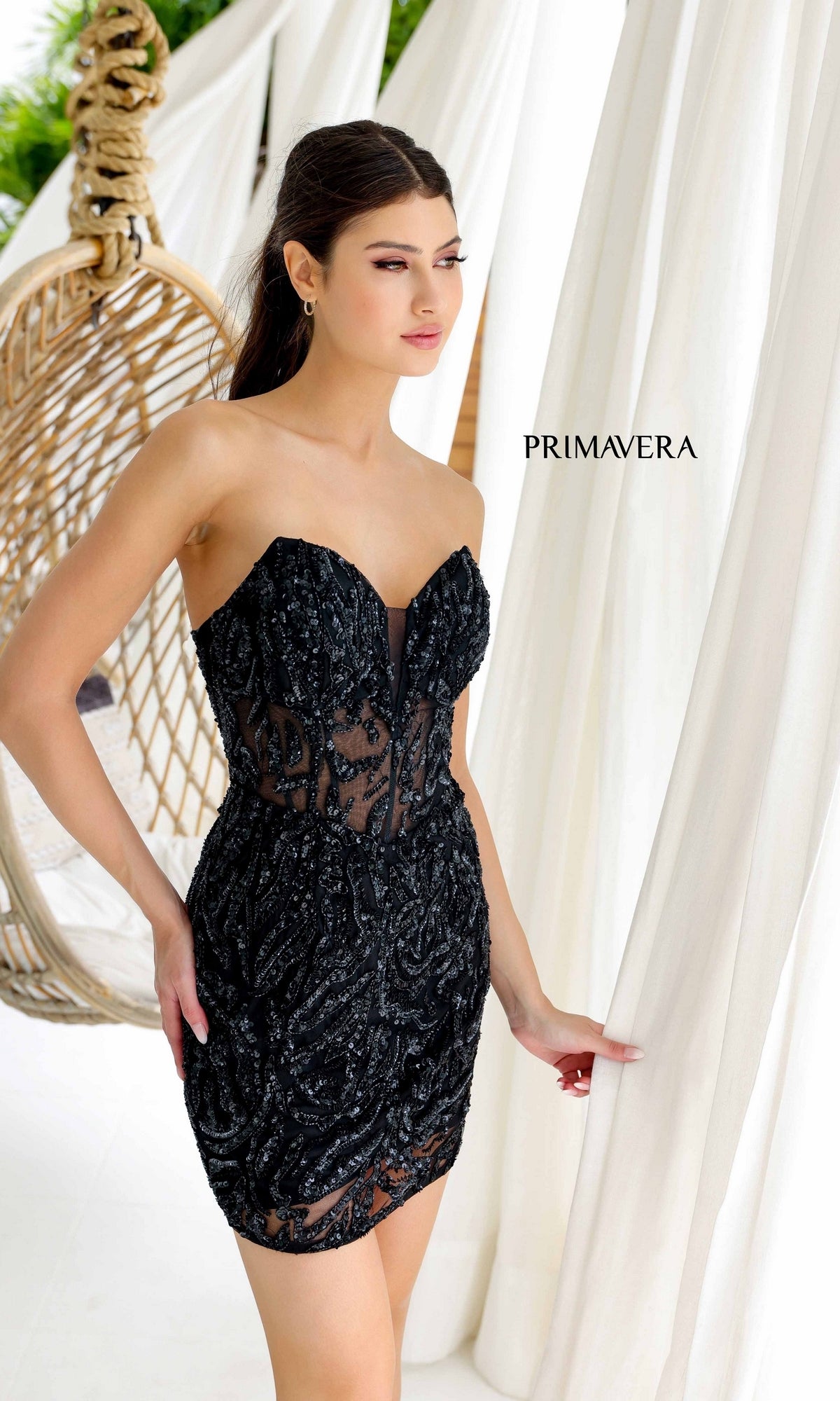 A woman stands by white drapes in the Short Homecoming Dress 4222 by Primavera—a black beaded strapless mini dress with sheer panels. A hanging rattan chair is in the background, and