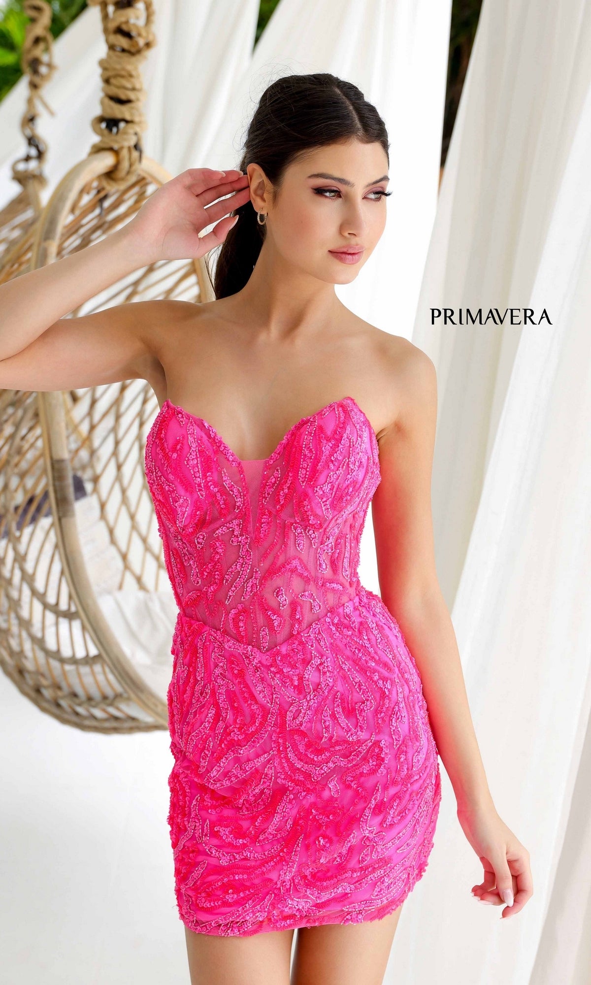 A woman poses in front of a hanging chair wearing the Short Homecoming Dress 4222 by Primavera, a bright pink strapless dress with sparkling sequins, with