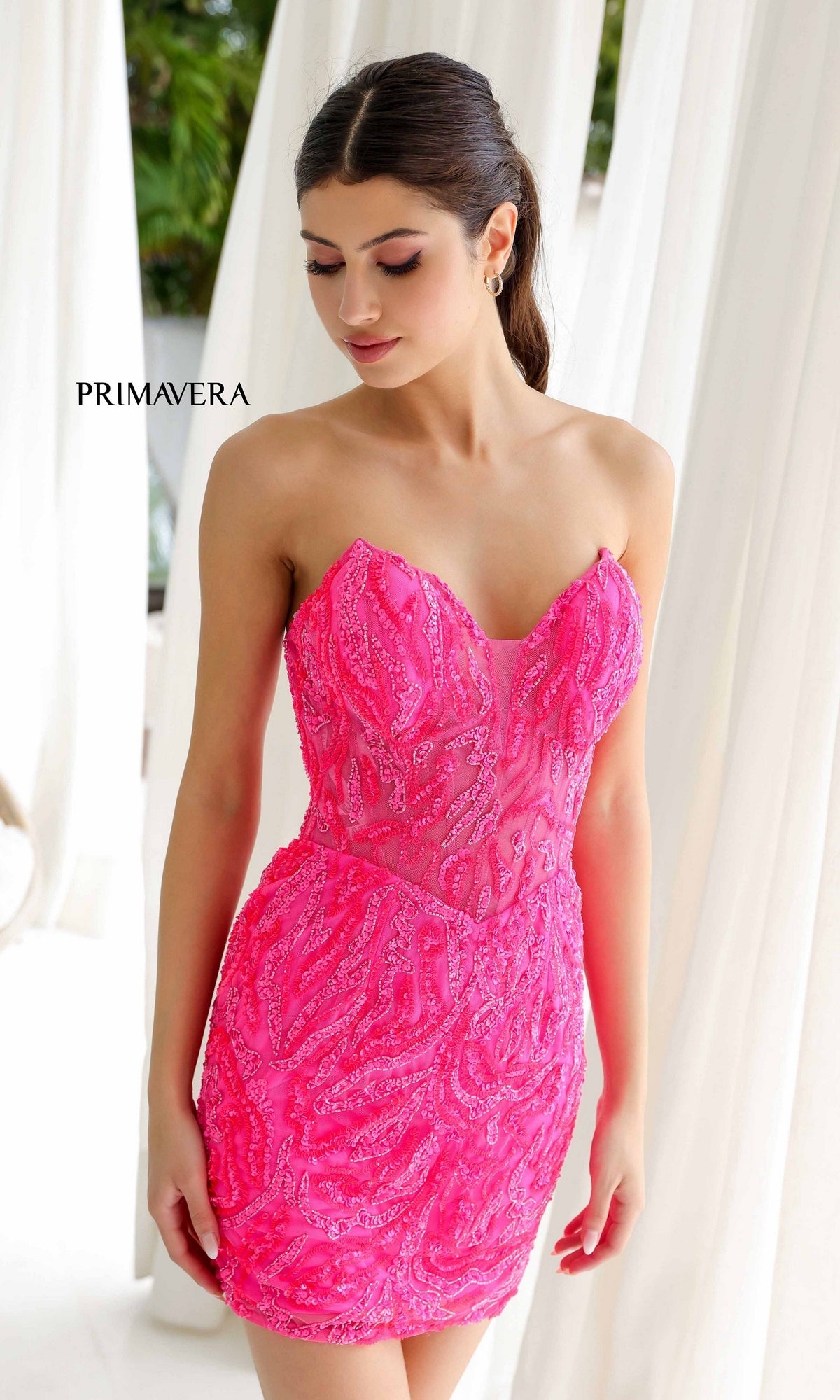 A woman with her hair styled back stands near white curtains, wearing the Short Homecoming Dress 4222 by Primavera featuring a strapless corset bodice and hot pink beaded mini skirt.