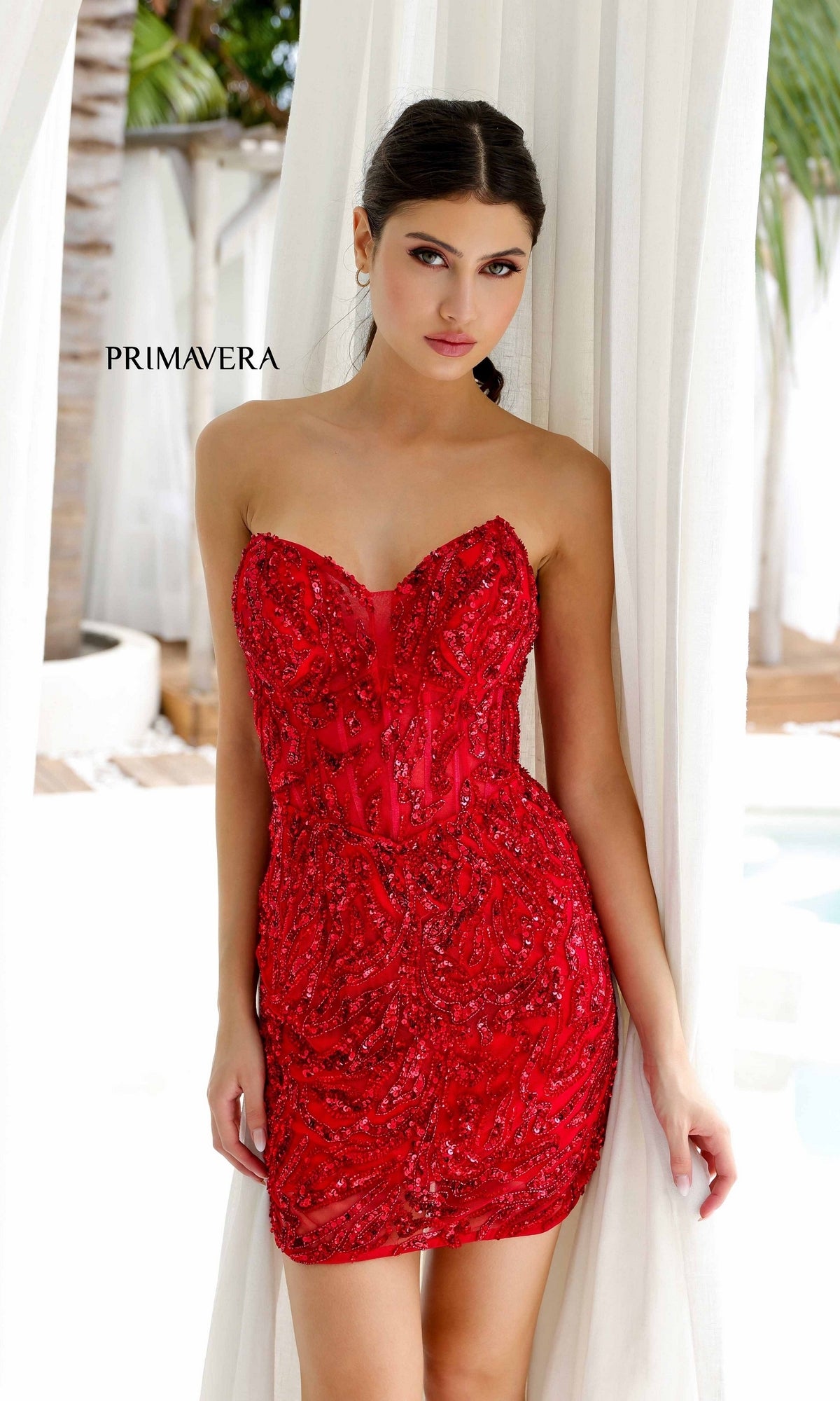 A woman stands between white curtains in front of palm trees and a pool, wearing the strapless, fitted Short Homecoming Dress 4222 by Primavera with red beading and a corset bodice.