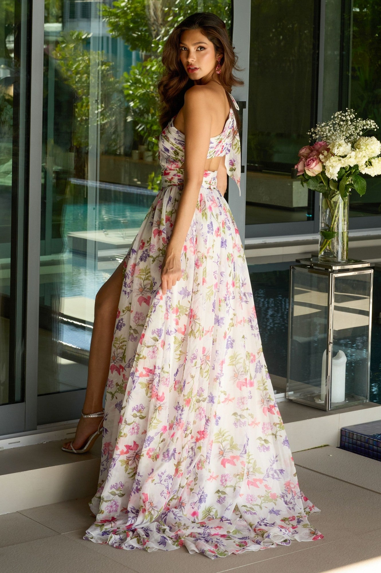 A woman in the Ava Presley 42237 long prom dress, featuring floral chiffon and a high slit, stands by a glass wall and looks over her shoulder with flowers and greenery in the background.