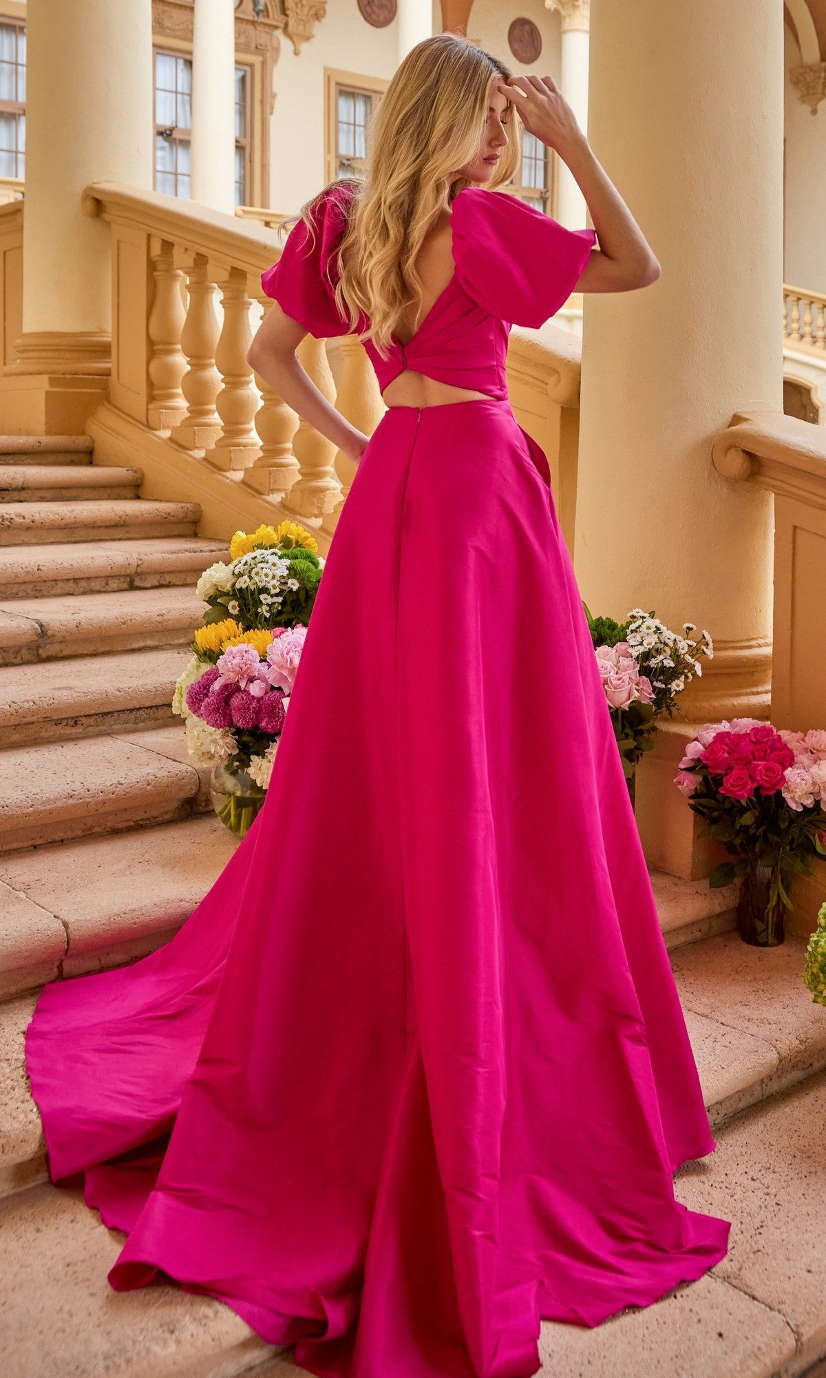 A woman wearing the Ava Presley 42269 puff-sleeve long prom dress stands on stone steps among flower arrangements, looking away from the camera.