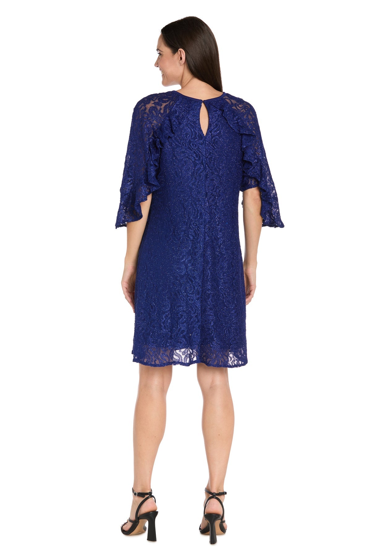 A woman stands facing away from the camera, wearing the R&M Richards 4226 short royal blue glitter lace dress featuring a knee-length a-line silhouette, flutter sleeves, keyhole back, paired with black heeled sandals.