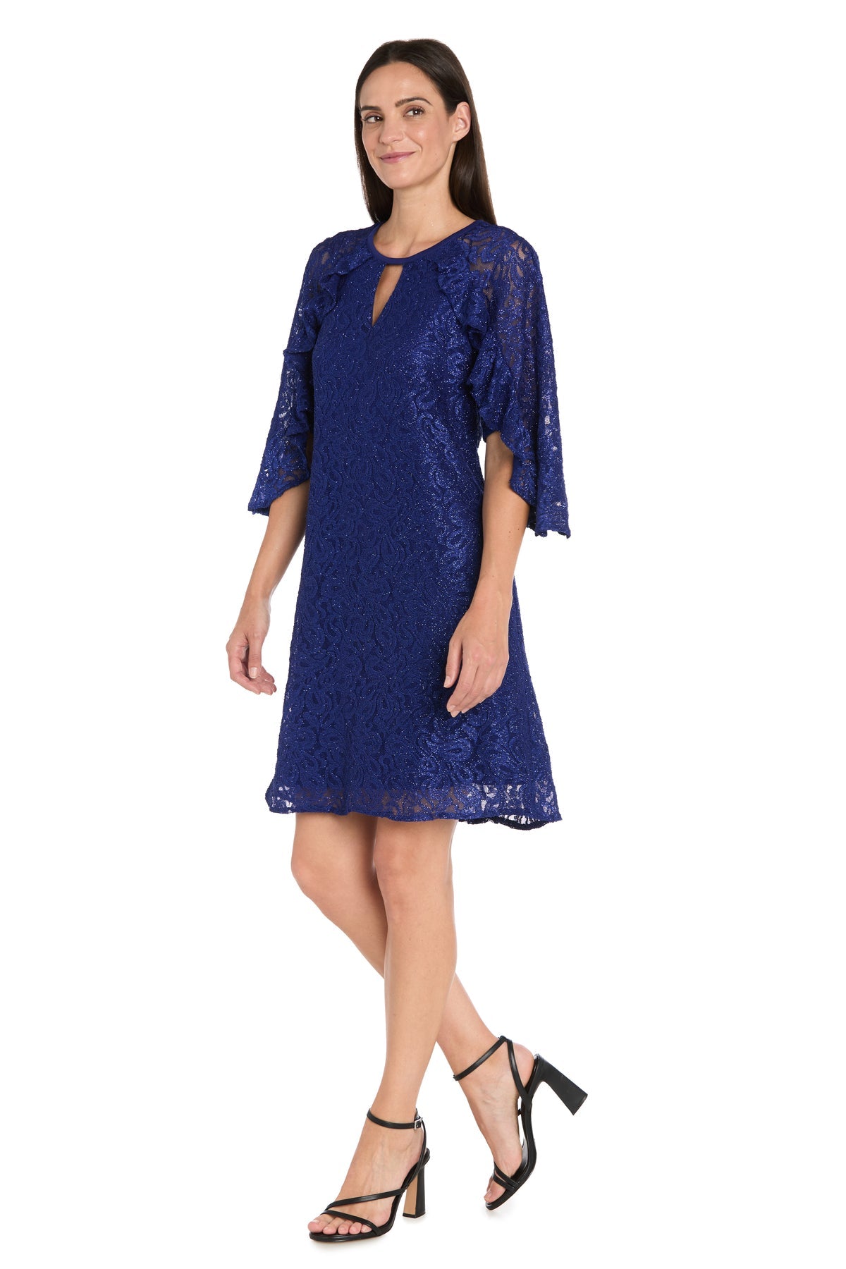 A woman models the R&M Richards 4226 Short Lace Semi-Formal Dress, a knee-length cobalt blue glitter lace A-line with elbow-length bell sleeves, paired with black heeled sandals, against a plain white background.
