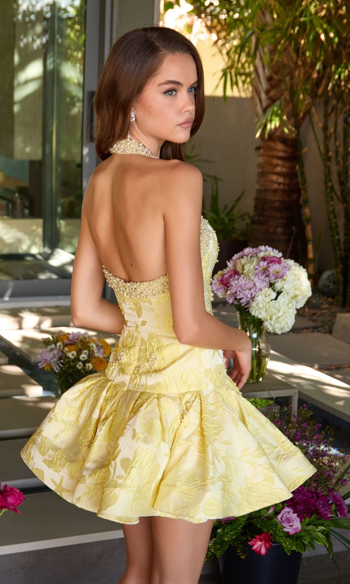 A woman in the Ava Presley 42399 Short Cocktail Dress stands indoors near glass doors, glancing over her shoulder. Flower arrangements are nearby and greenery is visible outside.
