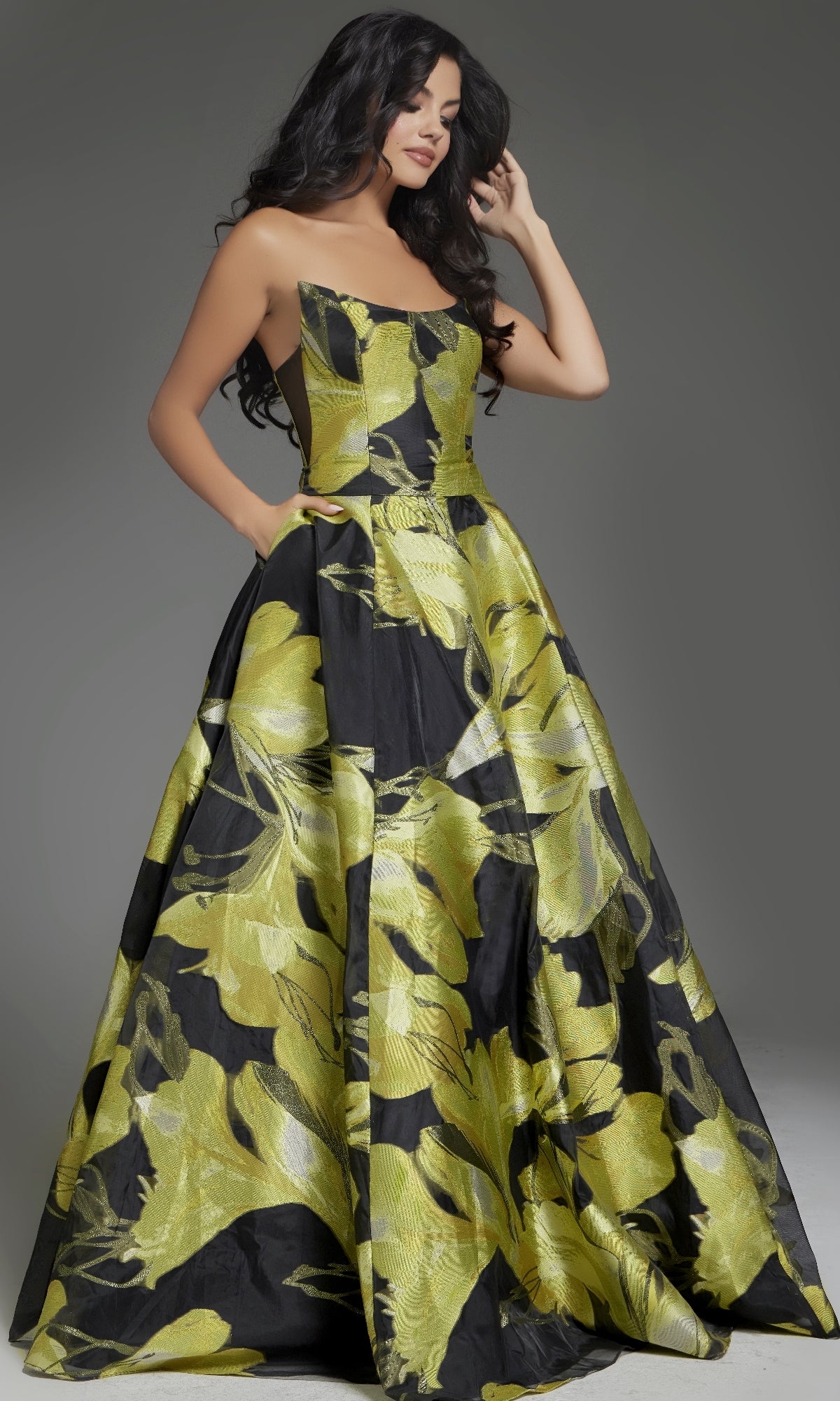A woman models the strapless Jovani 42435 long prom dress, featuring a bold yellow and black floral design, posed against a gray background—an elegant ball gown ideal for prom or special events.