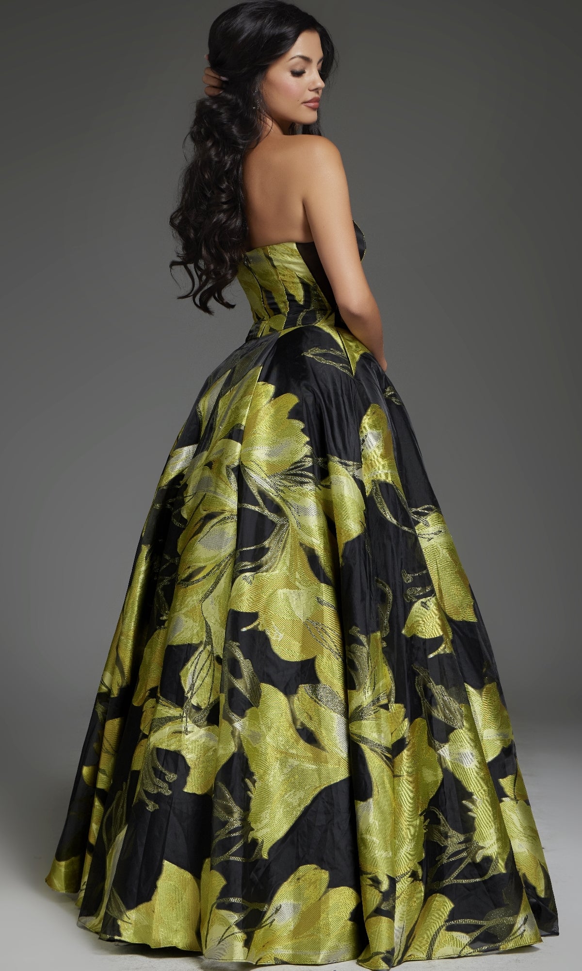 A woman poses with her back turned and head to the side, wearing the Jovani 42435 long prom dress—a strapless gown featuring bold yellow floral patterns—set against a neutral background.