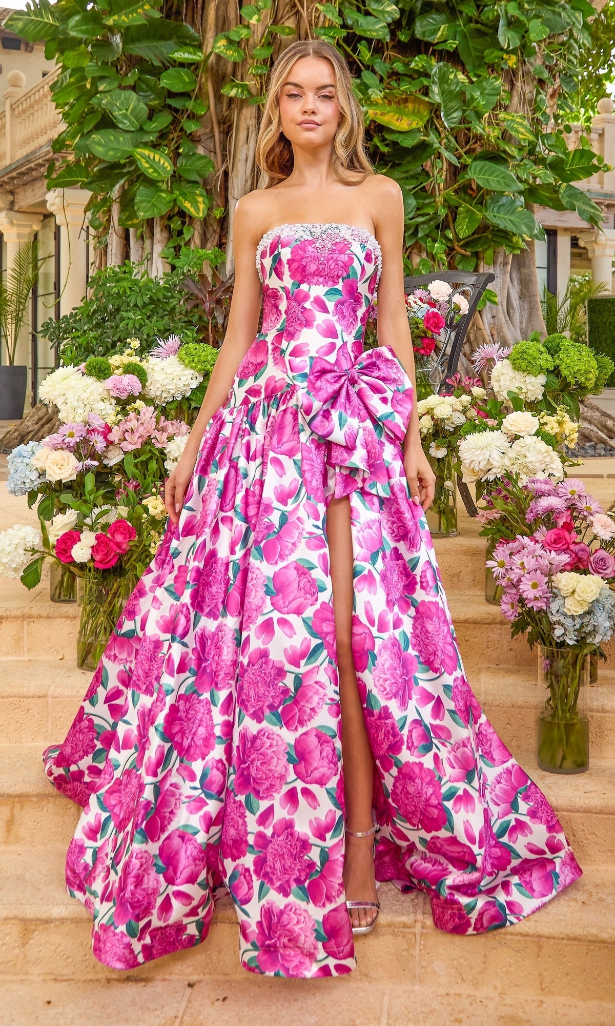 A vibrant pink strapless, floor-length Ava Presley 42459 floral prom dress with a side bow and high leg slit, modeled on stone steps among flower arrangements.