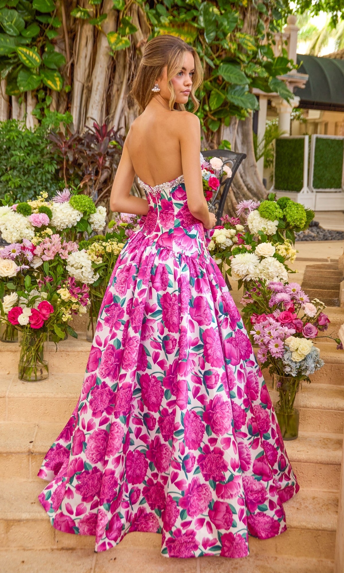 A person stands on stone steps wearing an Ava Presley 42459 floral, strapless, floor-length ball gown, surrounded by large bouquets of flowers.