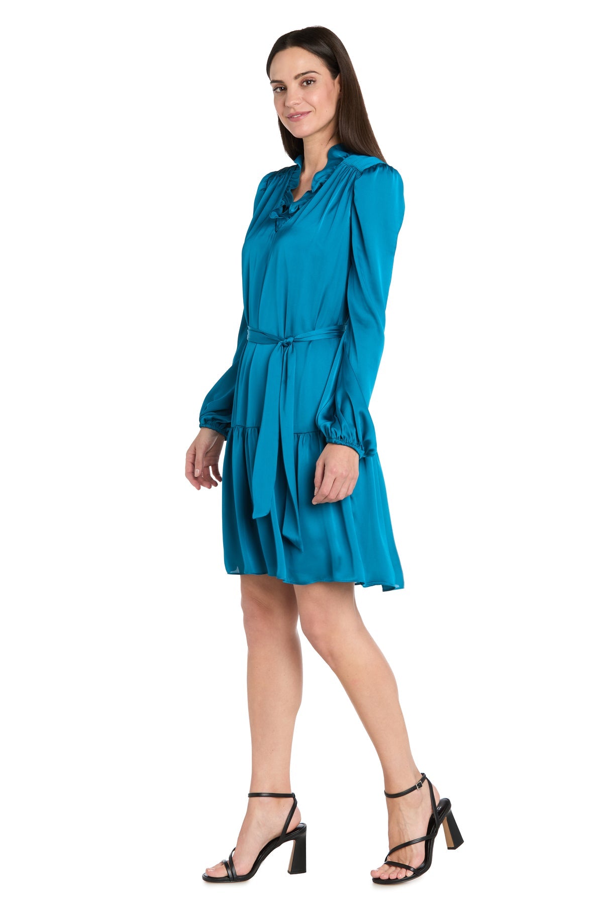A woman stands smiling against a white background, wearing the R&M Richards 4246P Petite Short Teal Party Dress with long sleeves, belted waist, ruffled hem, and black strappy heels.