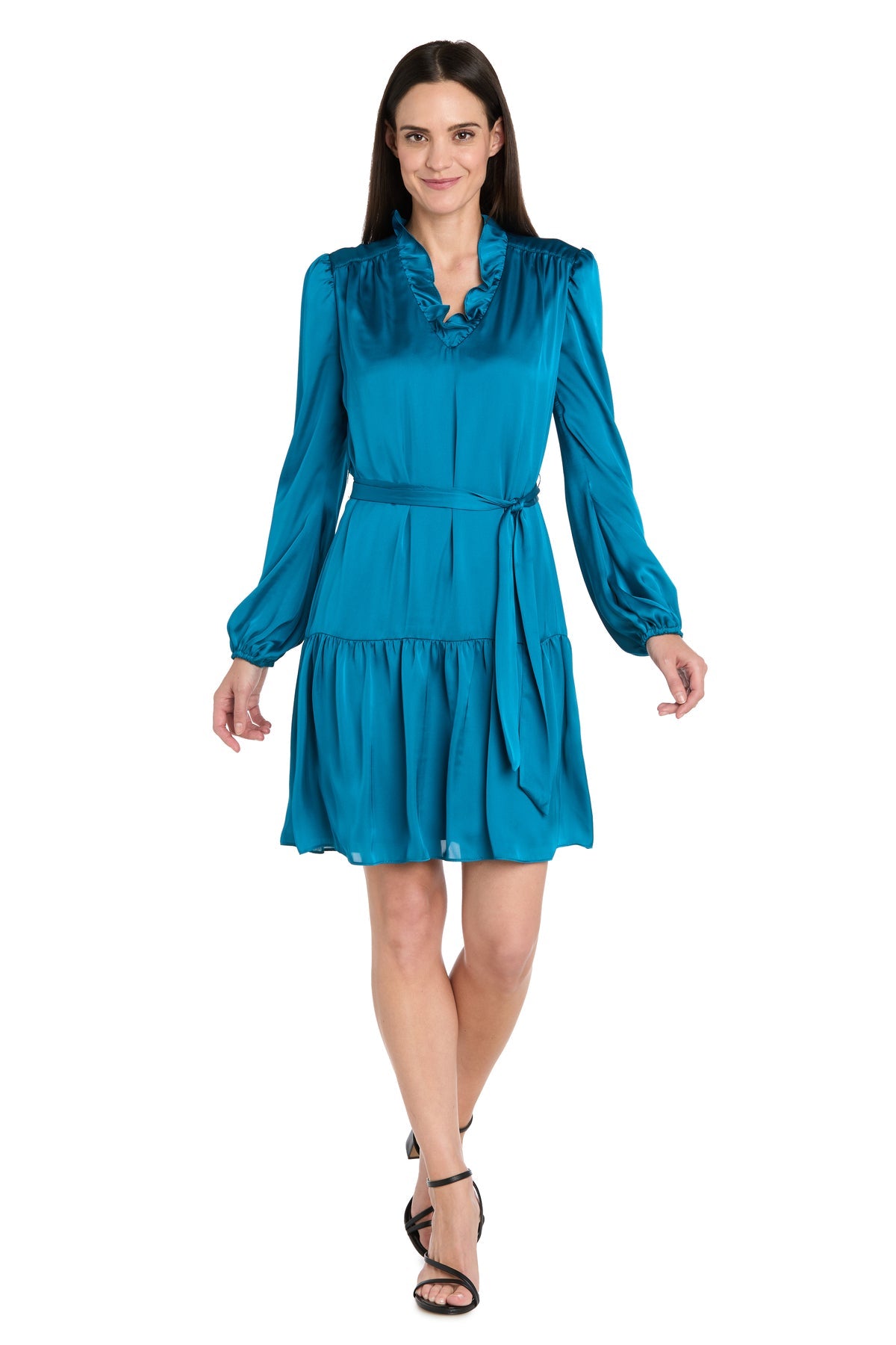A woman walks forward and smiles in a Petite Short Teal Party Dress (R&M Richards 4246P) with long sleeves, a belted waist, and a ruffled collar, set against a white background.