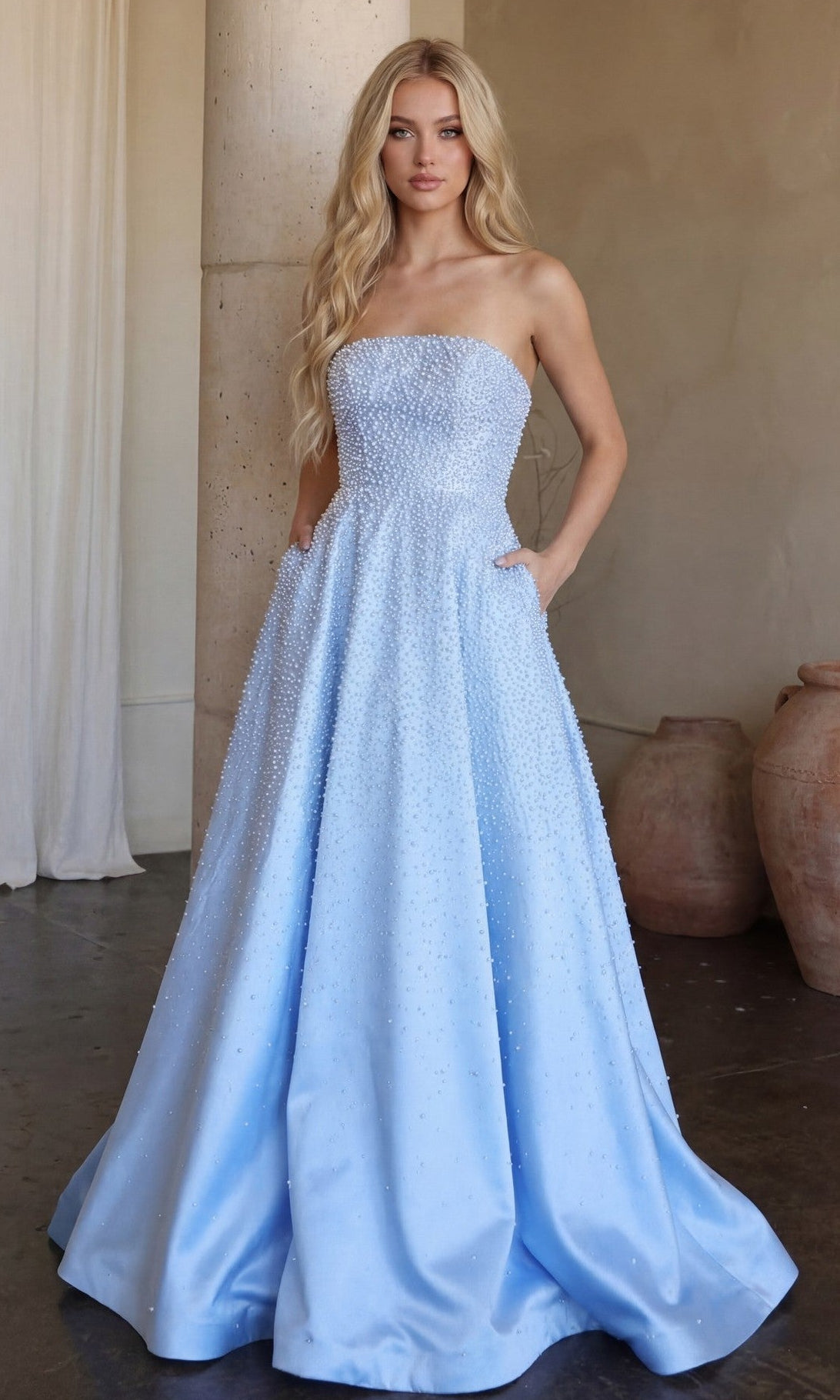 A woman wearing the Ava Presley 42500 Strapless Long Pearl Prom Dress in Powder Blue stands indoors near a column and pottery.