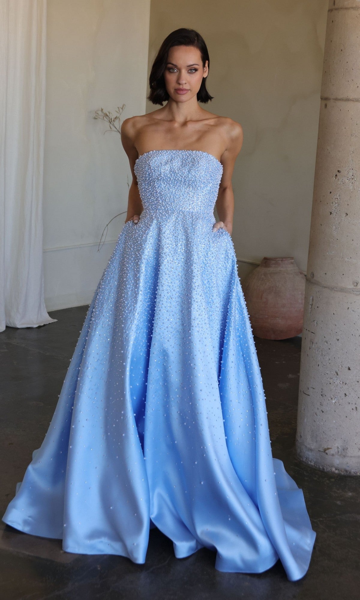 A woman stands indoors wearing the Ava Presley 42500, a long, strapless, light blue prom dress adorned with pearls and featuring an elegant A-line silhouette.