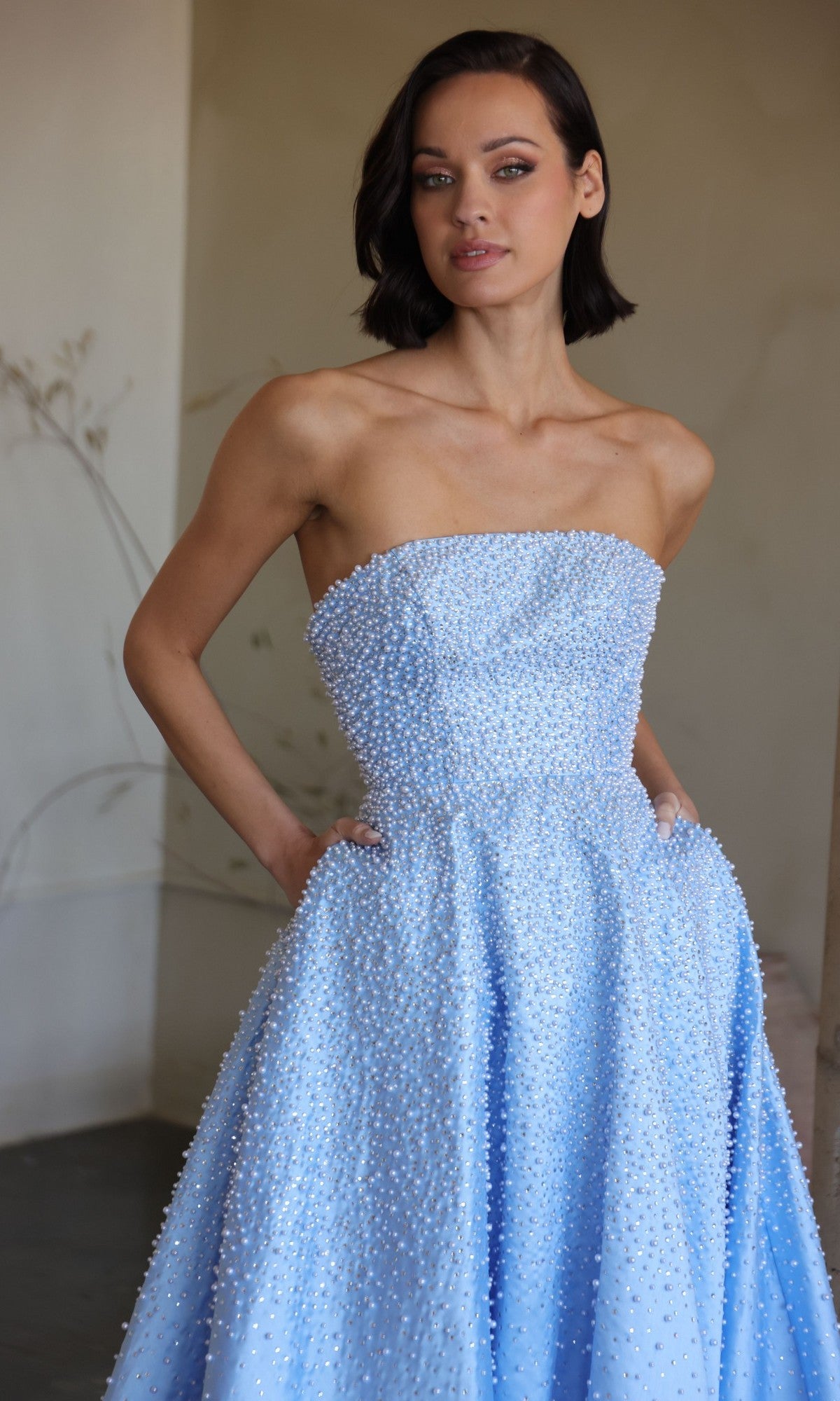 A woman wearing the Ava Presley 42500, a strapless light blue long prom dress with small white beads and pockets in the A-line skirt, stands indoors looking at the camera.