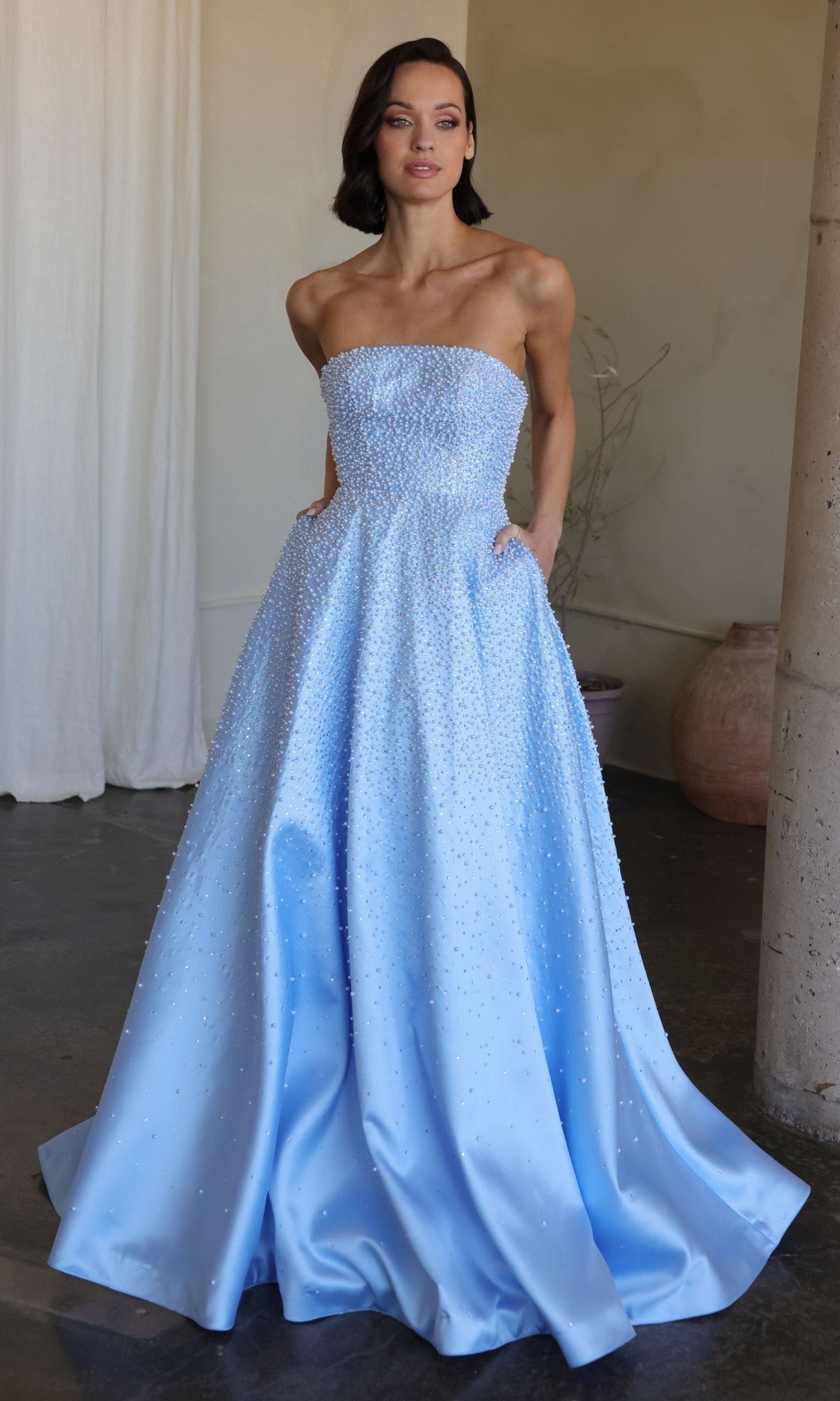 A woman stands indoors wearing the Ava Presley 42500, a powder blue, strapless, floor-length prom dress adorned with small beads—perfect for special occasions.