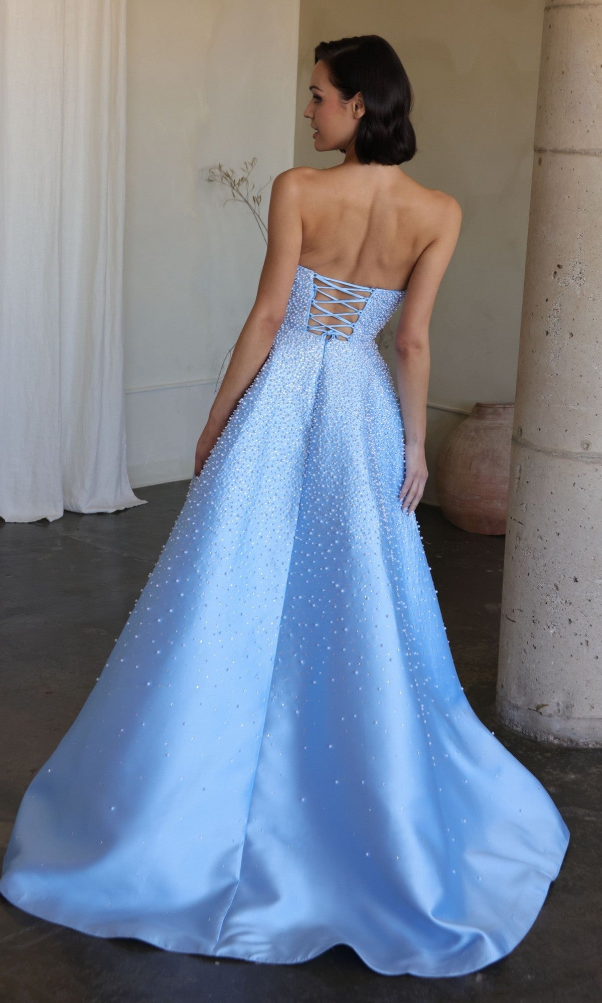 A woman in the Ava Presley 42500 long prom dress—a light blue, strapless, floor-length gown with a lace-up back and beaded details—stands facing away in an elegant indoor setting.