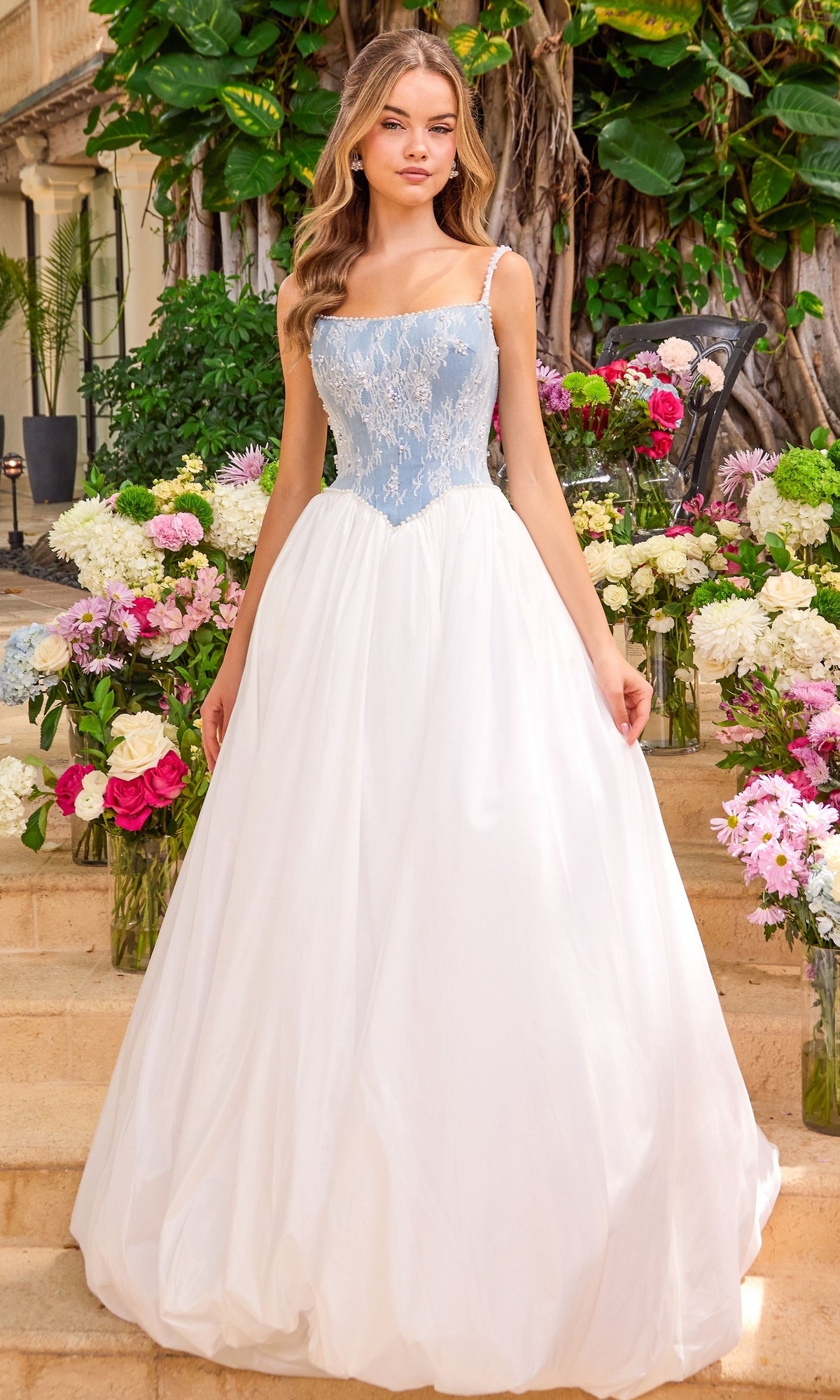 A person stands on stone steps among floral arrangements wearing an Ava Presley 42519 ball gown with a light blue, lace-appliqued bodice and a floor-length white skirt.