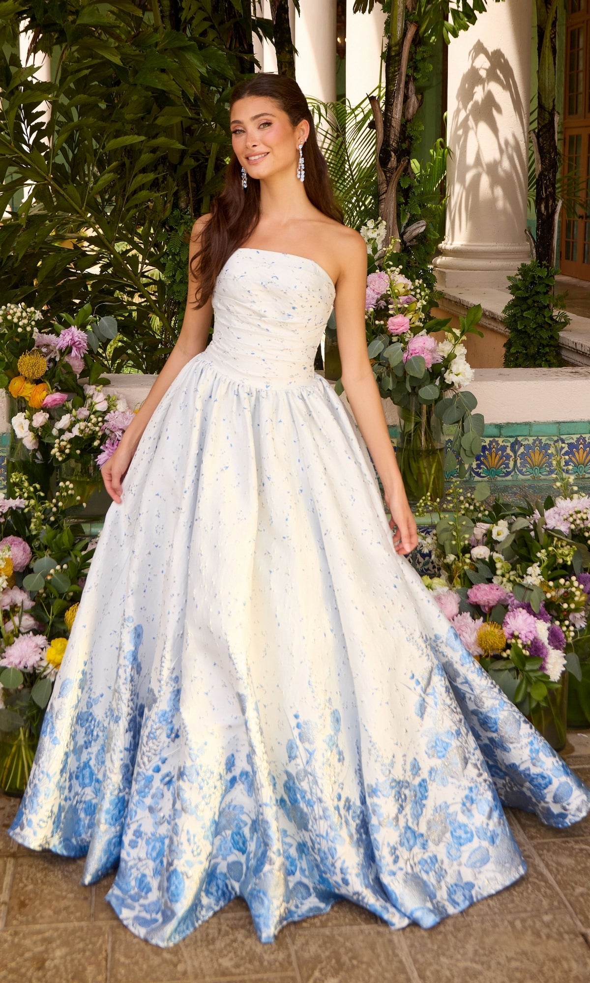 A woman wears a Long Prom Dress: Ava Presley 42540, a strapless, floor-length ballgown with a white bodice and a blue floral ombre skirt, standing in front of floral arrangements.