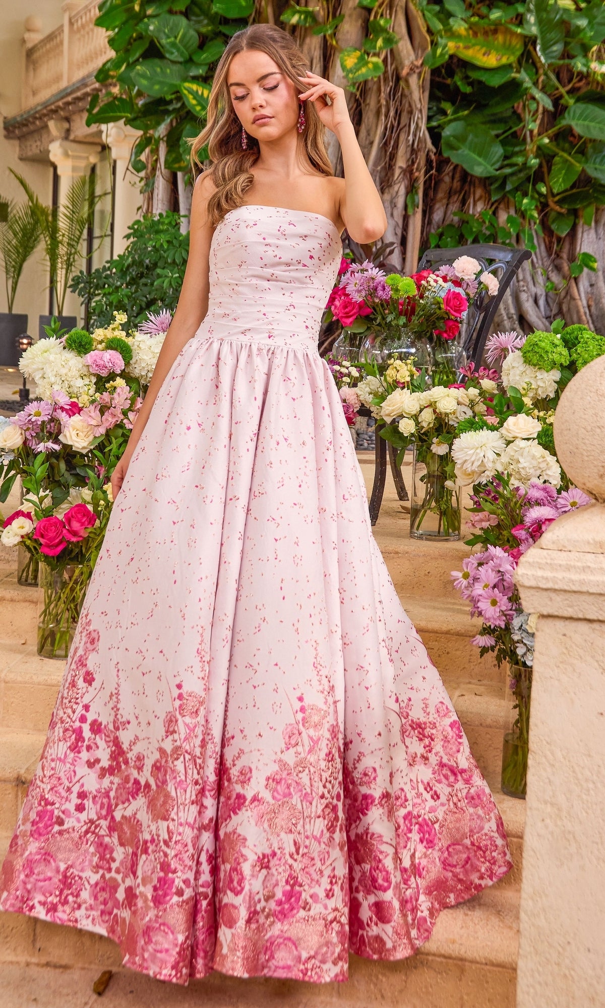A person wearing a pink Ava Presley 42540 strapless floral prom ball gown that transitions into a dense dark pink print at the hem, standing on stone steps surrounded by bouquets of flowers.