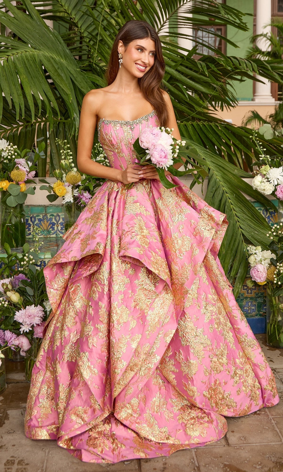 A woman wears a pink, strapless Ava Presley 42541 ballgown with gold floral patterns and a beaded neckline while holding a bouquet of pink peonies.