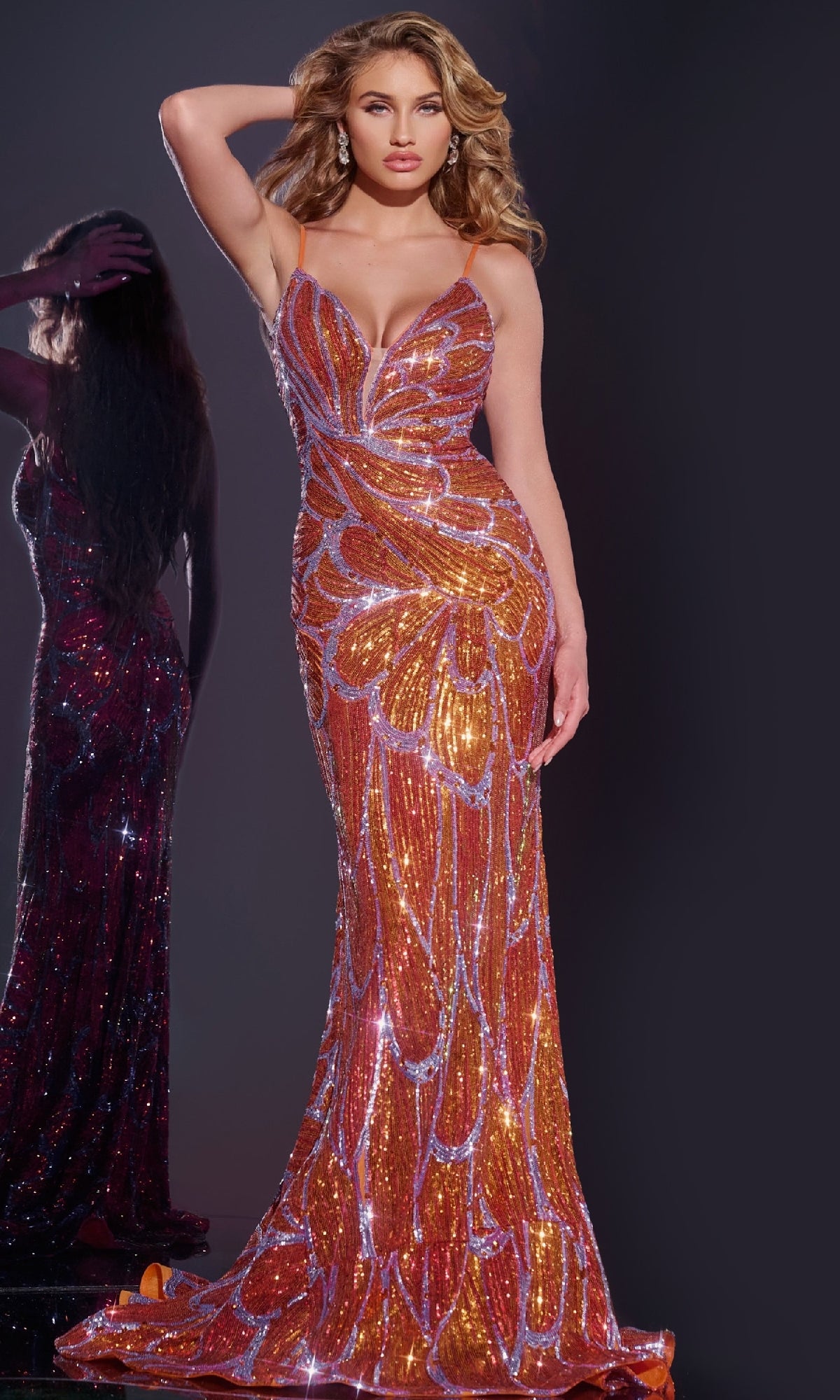 A woman in a fitted, floor-length orange/lilac Jovani 42600 Deep V-Back Long Sequin Prom Dress with spaghetti straps stands against a dark background, one hand in her hair. Another woman appears in the background.