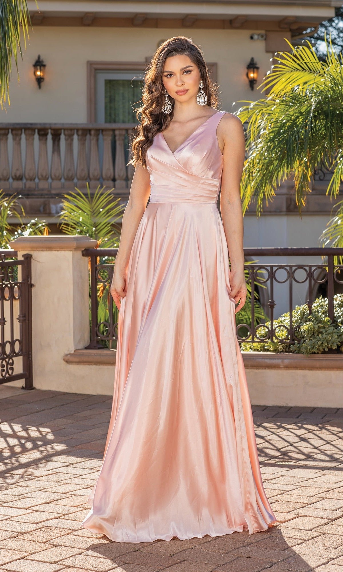 A woman stands outdoors on a paved patio, surrounded by greenery and a building in the background, wearing the Wrap-Bodice Long A-Line Prom Dress 4262—a long, shiny blush pink evening gown.