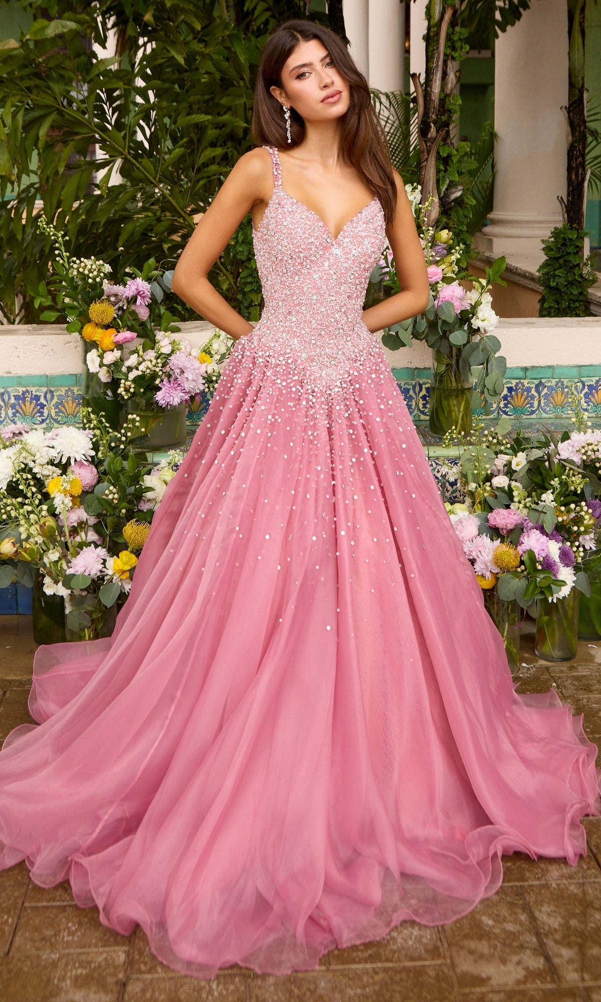 A woman wearing a blush pink Ava Presley 42643 beaded prom dress with a floor-length ball gown skirt, standing in front of floral arrangements.