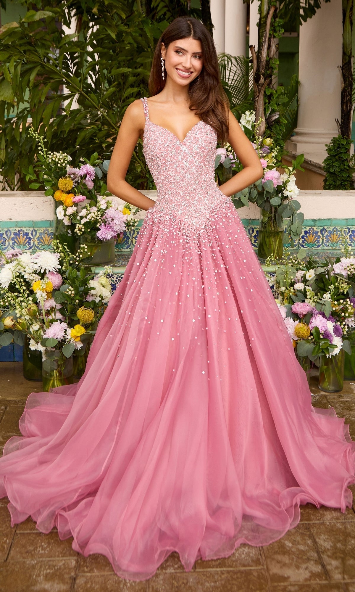 A woman stands in front of potted flowers wearing a floor-length pink Long Prom Dress: Ava Presley 42643 with a beaded bodice and flowing skirt.