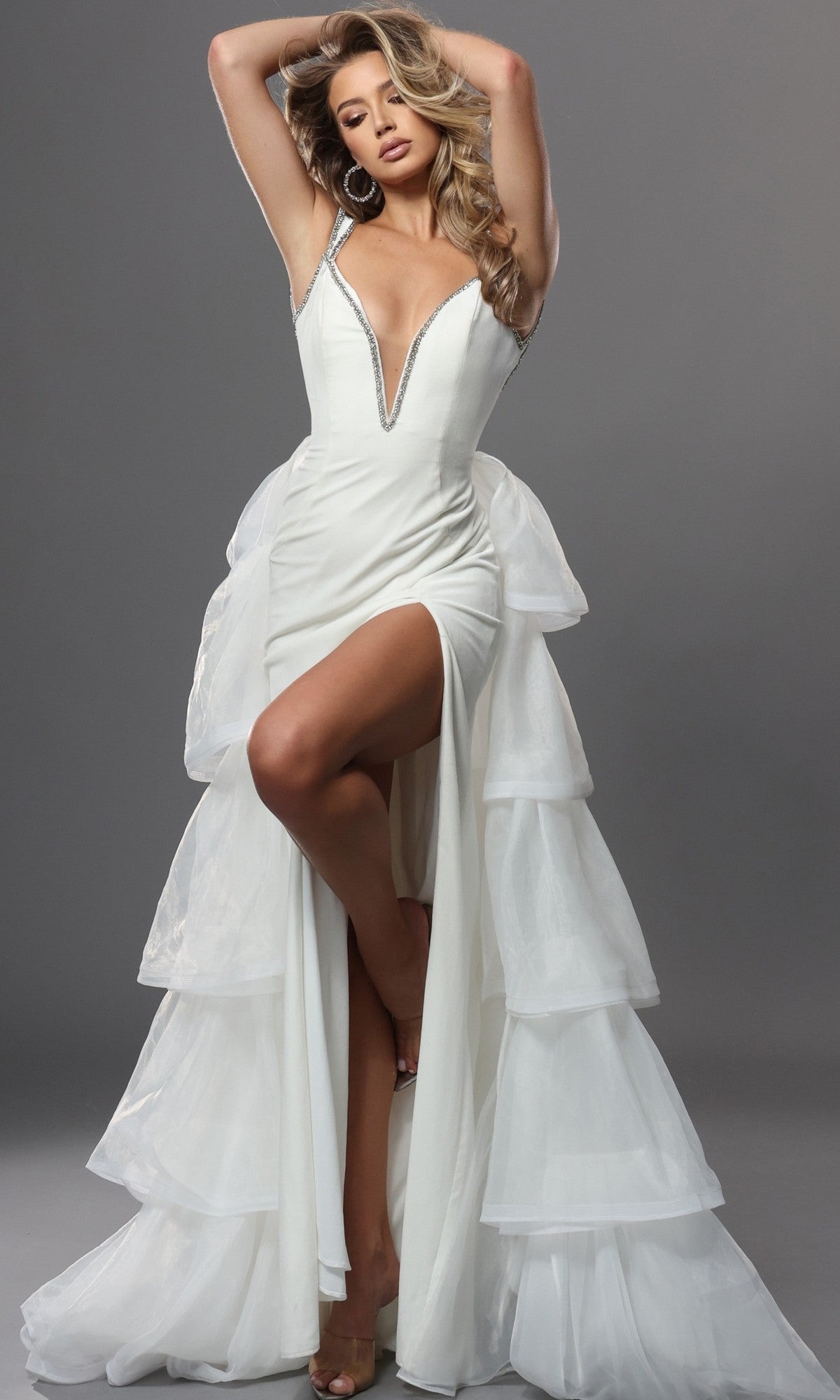 An elegant woman poses with arms raised against a gray background, wearing the off-white Jovani 42661 long formal dress featuring a sleeveless design, deep V neckline, tiered tulle ruffles, high slit, and shimmering silver trim.