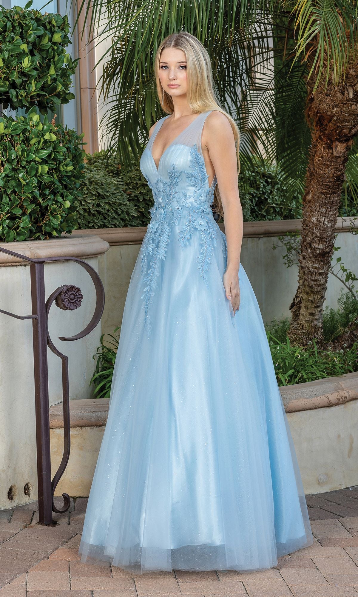 A young woman stands outdoors in the Backless Glitter Tulle Ball Gown Prom Dress 4272—a long, light blue, sleeveless gown with delicate detailing—surrounded by greenery and stonework.