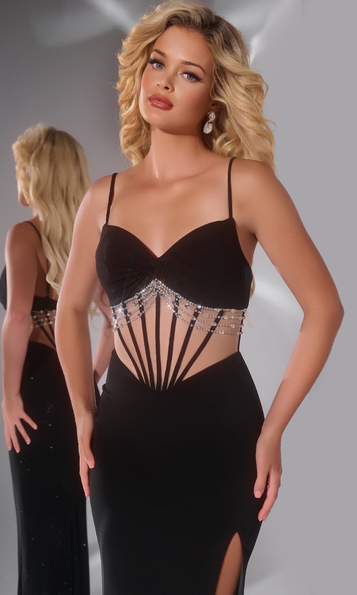 A woman poses in the Long Prom Dress: Jovani 42758, a black evening gown featuring sheer cutouts, crystal embellishments, and a sweetheart neckline. Her reflection appears in a mirror against a neutral background.