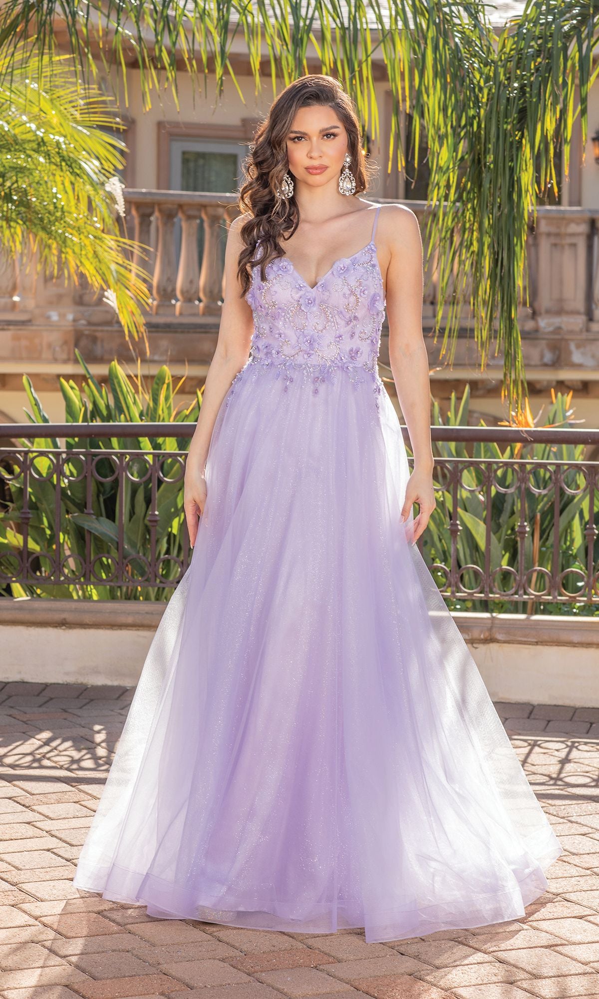 A woman stands outdoors in the Long A-Line Tulle Prom Ball Gown with Flowers 4276, featuring 3-D floral designs, thin straps, and a flowing skirt. Lush greenery and a building appear in the background.