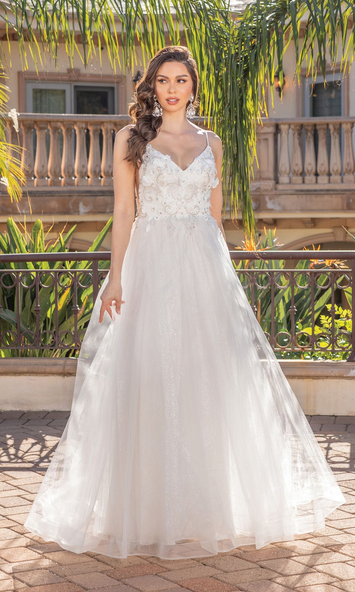 A woman stands outdoors by a balcony and greenery wearing the Long A-Line Tulle Prom Ball Gown with Flowers 4276, featuring a white, floor-length silhouette and 3-D floral designs.