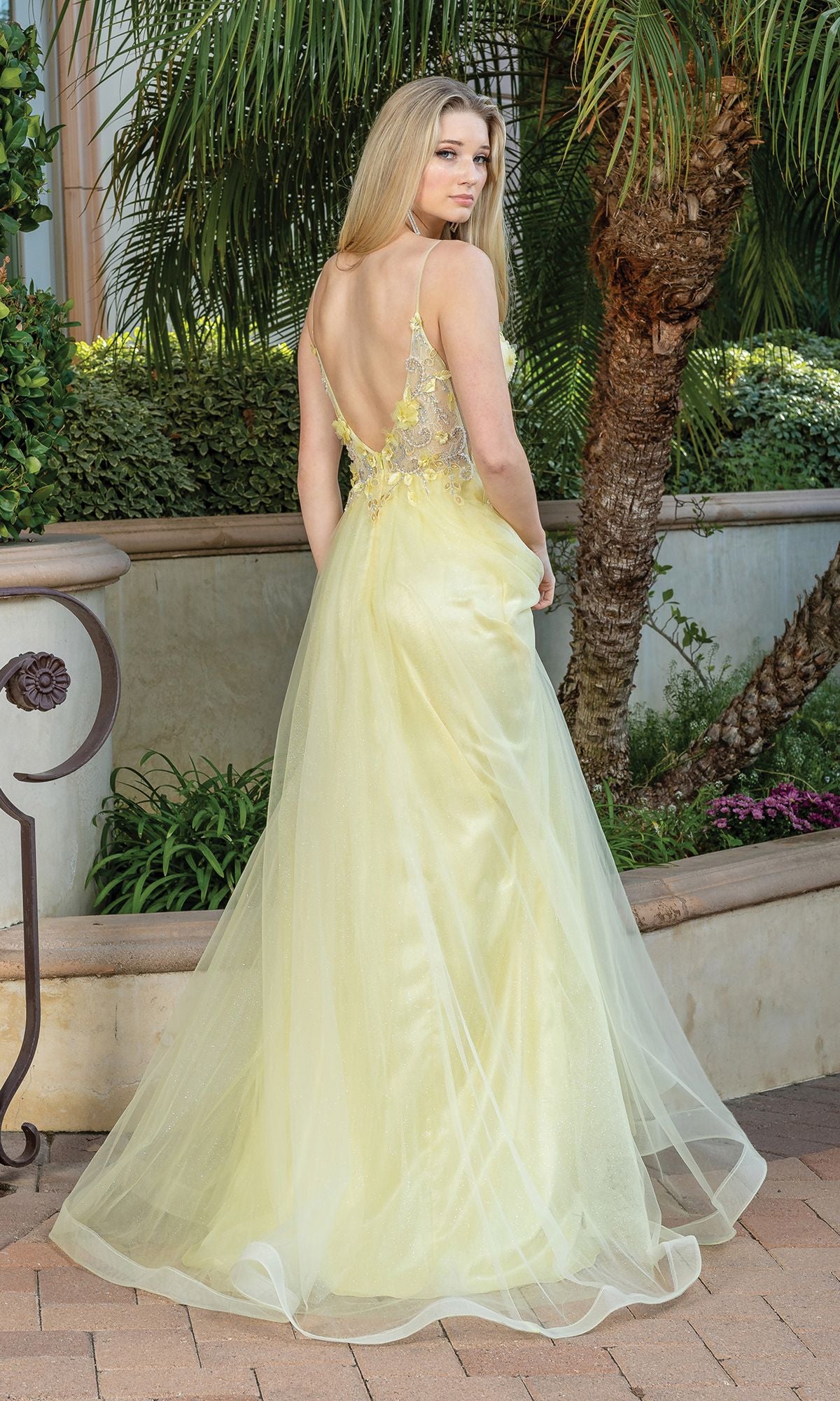 A woman wearing the Long A-Line Tulle Prom Ball Gown with Flowers 4276 stands outdoors near a palm tree, looking over her shoulder in a pale yellow gown with a sheer back.
