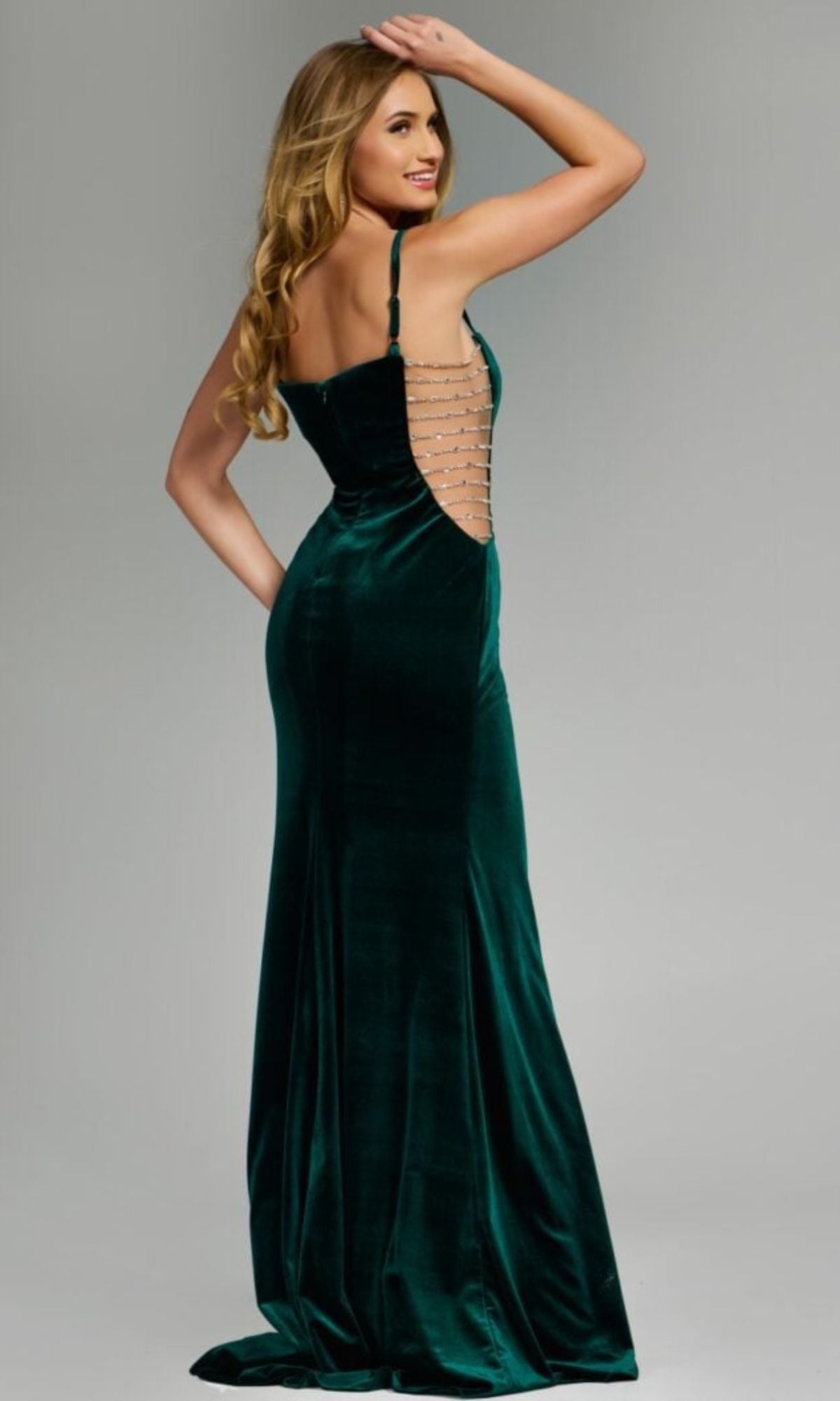A woman models the Jovani 42783 long prom dress in dark green velvet, showcasing its open back with horizontal straps and embellished sheer sides, while standing and looking over her shoulder against a neutral background.