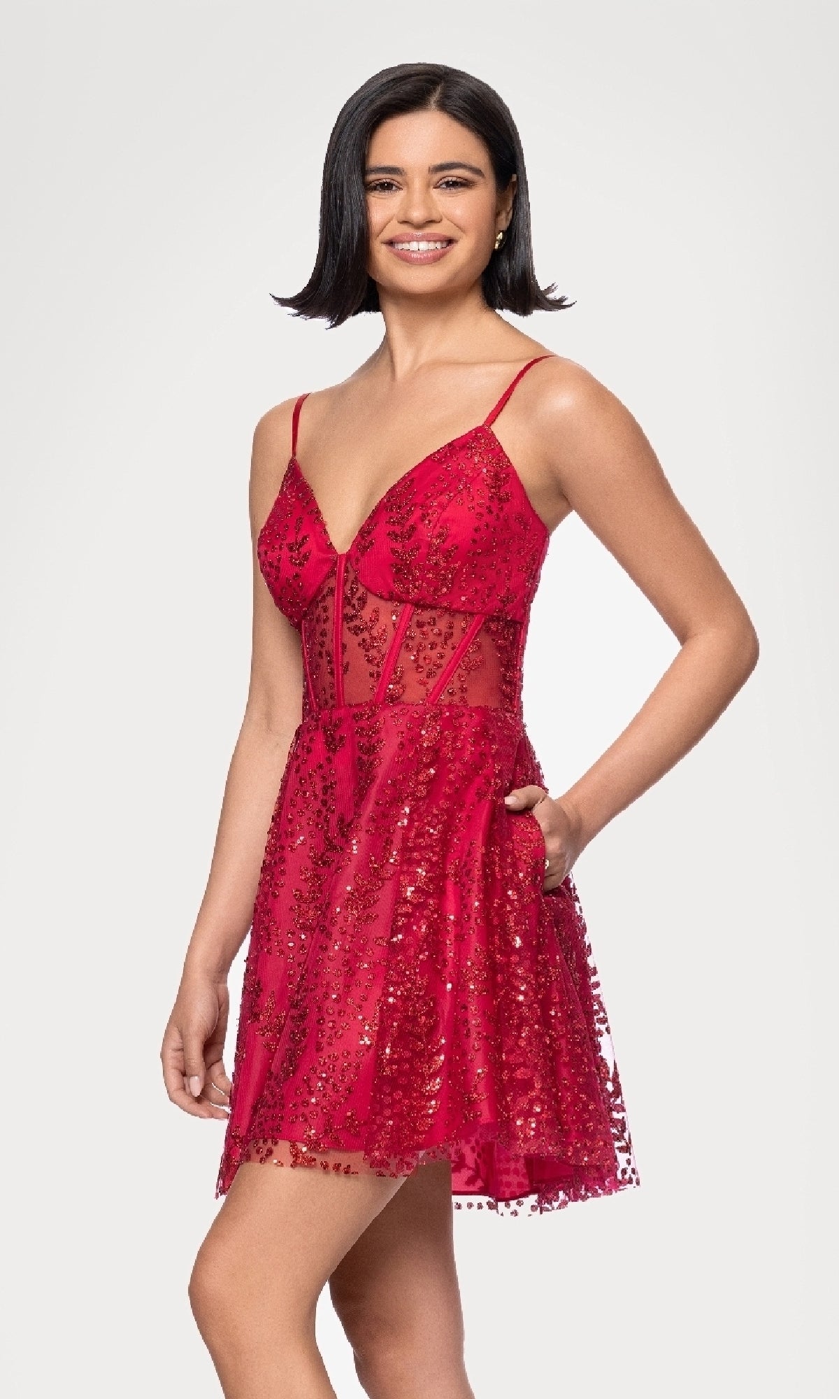 A woman smiles against a plain background in the Short Red Glitter Hoco Dress by Blondie Nites 4279BN, featuring thin straps, sheer paneling, and sparkling sequin embellishments.