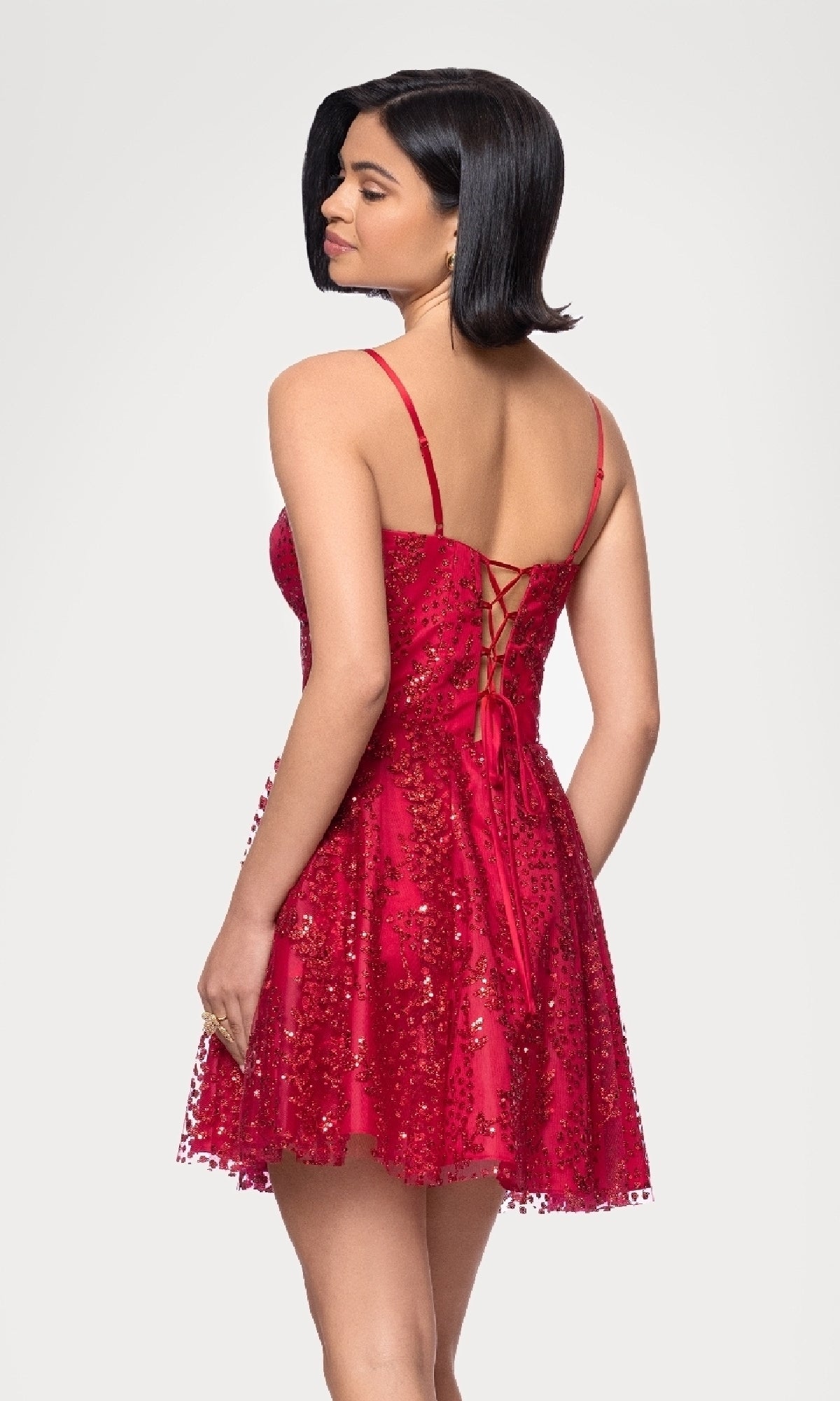 A woman, facing away from the camera against a plain background, wears the Short Red Glitter Hoco Dress: Blondie Nites 4279BN featuring thin straps, a lace-up back, and sequin embellishments.