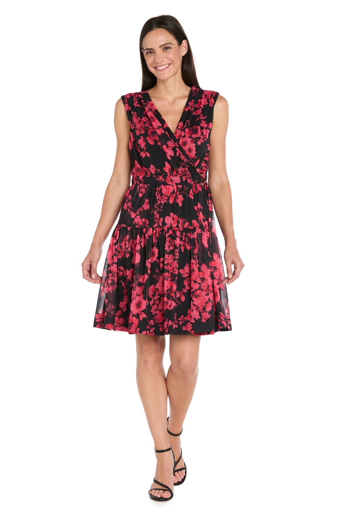 A woman stands and smiles against a white background, wearing the R&M Richards 4299 Red Print Wedding Guest Dress—a sleeveless black cocktail dress with a red floral pattern and V-neckline—paired with black strappy sandals.