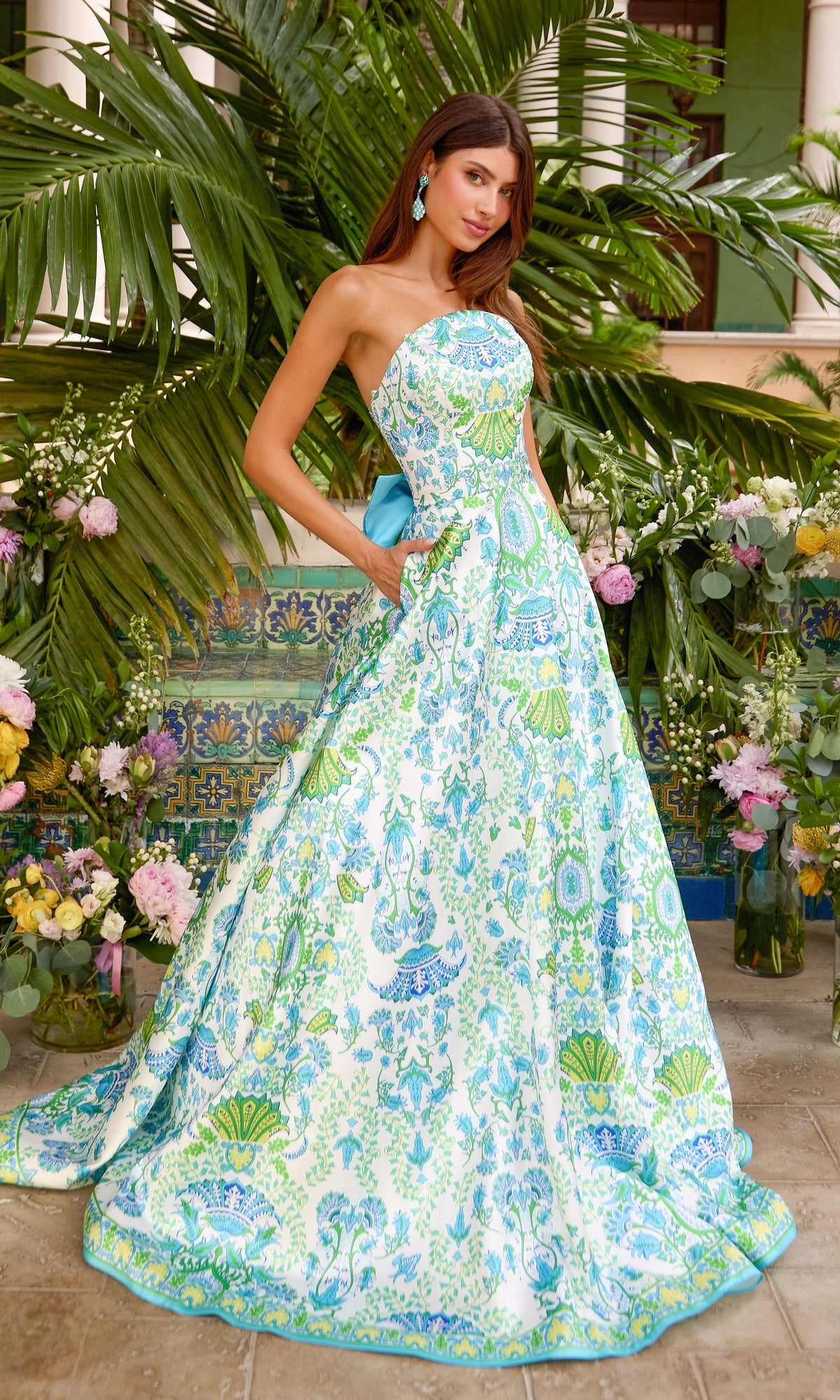 A woman wearing an Ava Presley 43017 strapless, floor-length white prom ball gown featuring a blue and green botanical print and a solid blue bow at the back.