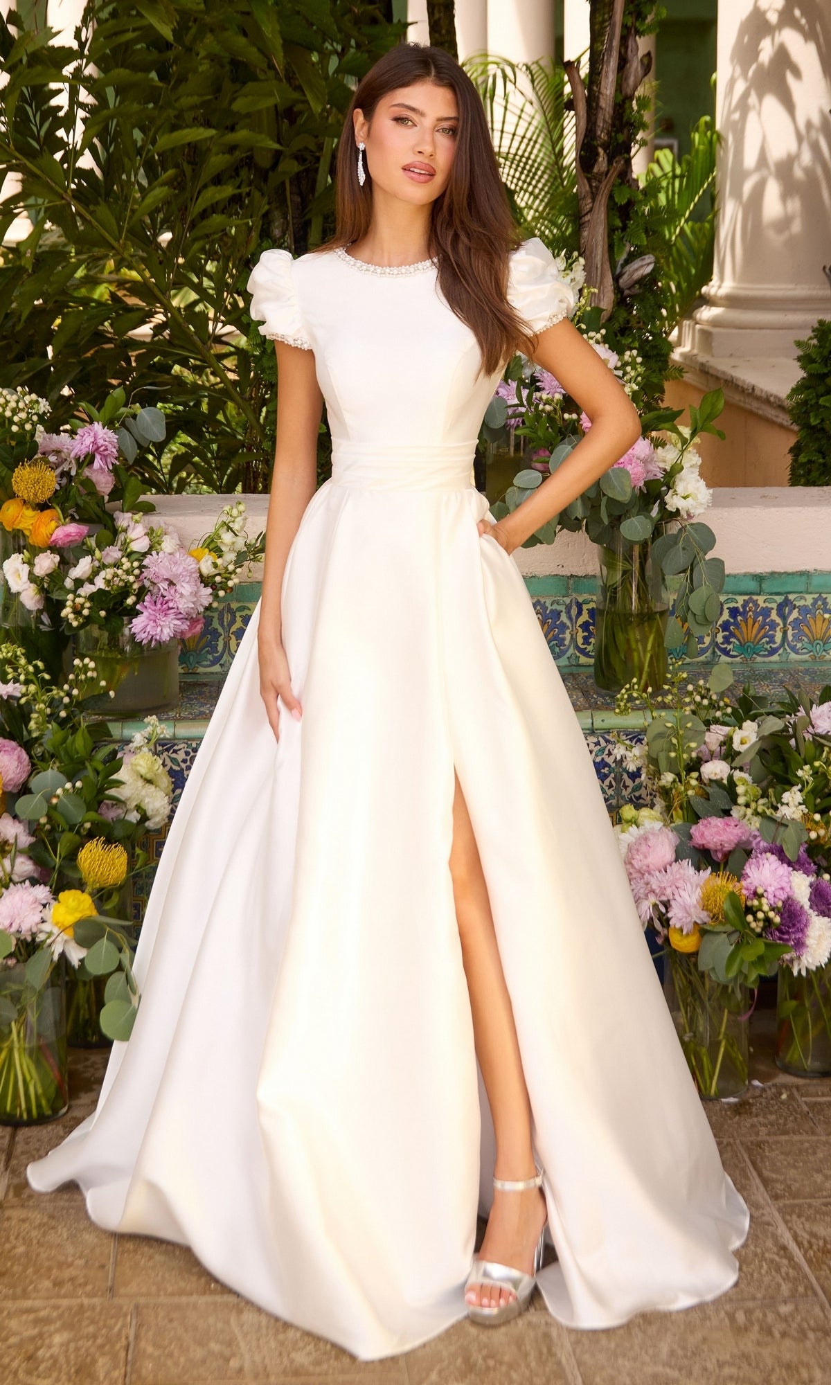 A woman in a white Ava Presley 43023 floor-length puff sleeve prom dress with a jewel neckline and high leg slit, standing among floral arrangements.