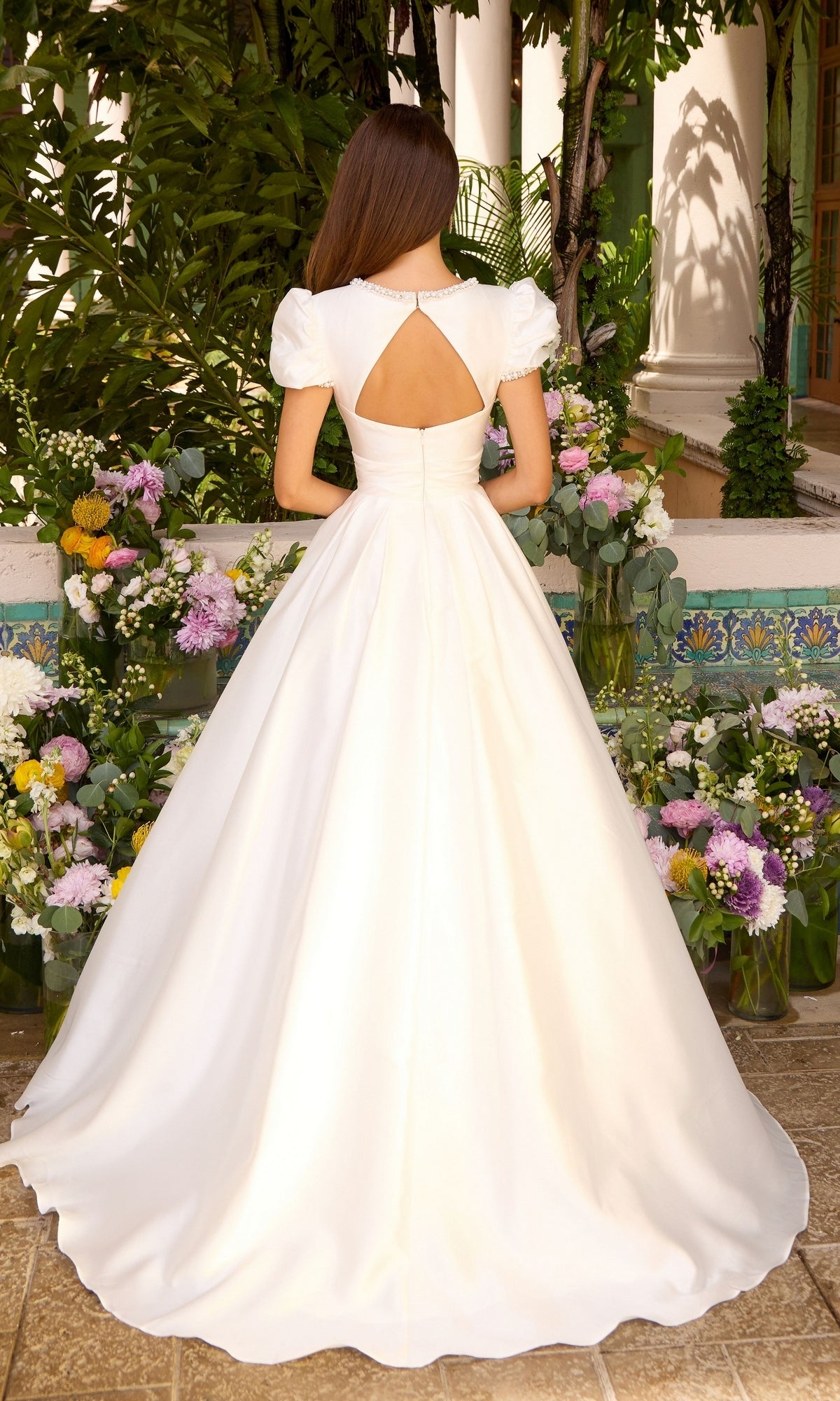 Back view of a white, floor-length Ava Presley 43023 prom dress with puff sleeves, a circular back cutout, and a full skirt, standing in front of floral arrangements.