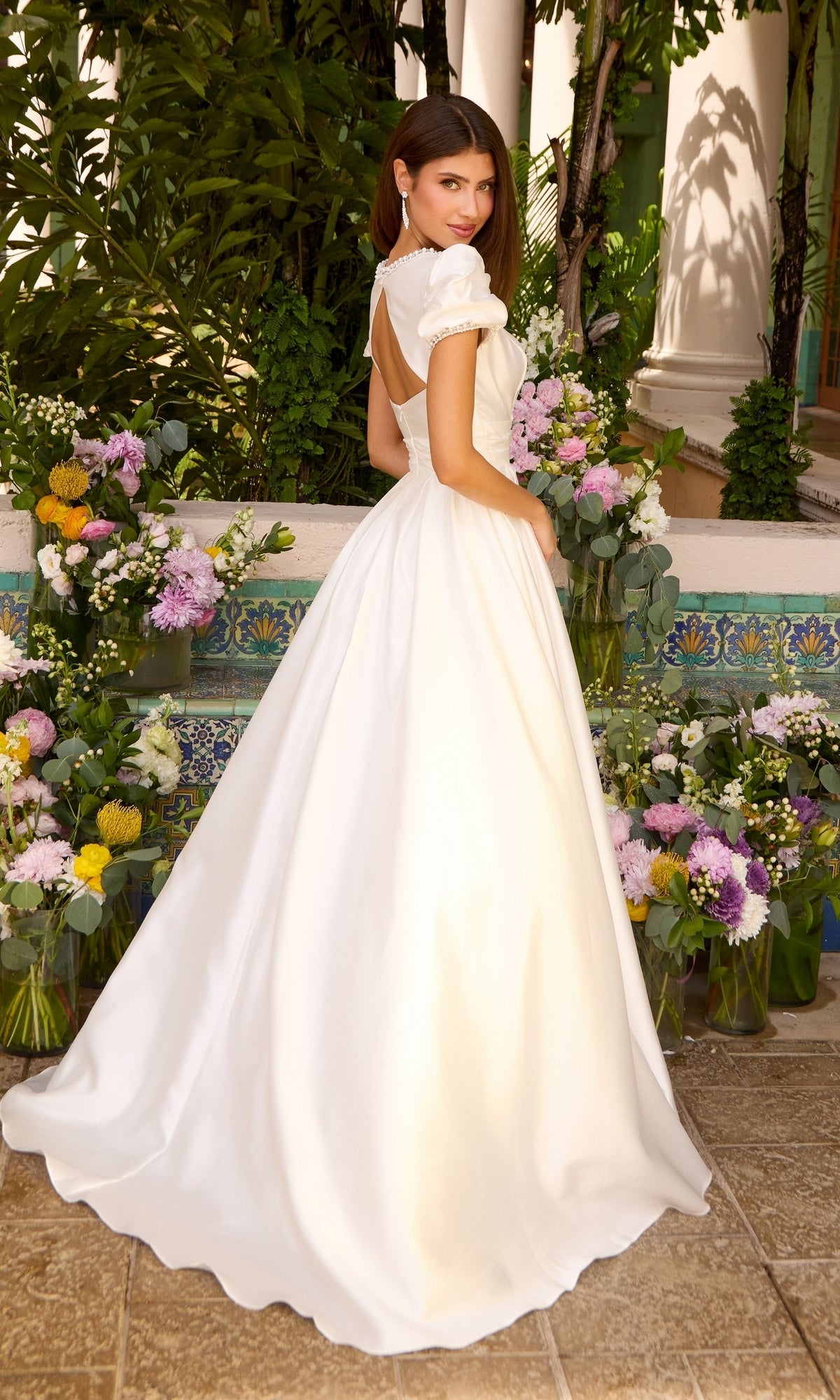 A person wearing a white, floor-length Ava Presley 43023 gown with puff sleeves and an open back, standing among arrangements of colorful flowers.