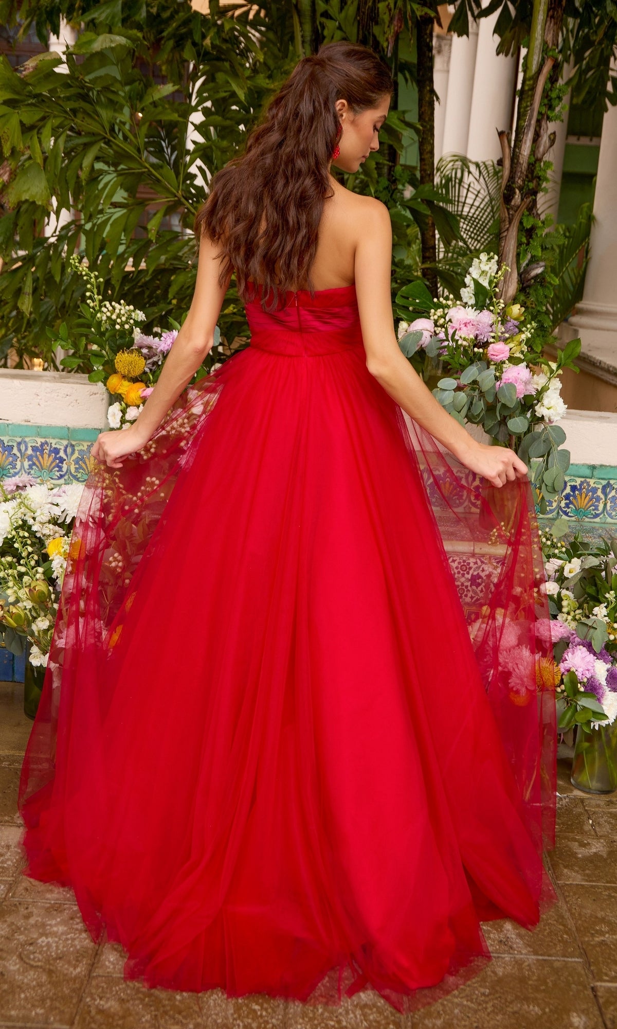 A person stands in a garden wearing an Ava Presley 43045 strapless, floor-length red gown with a full tulle skirt.