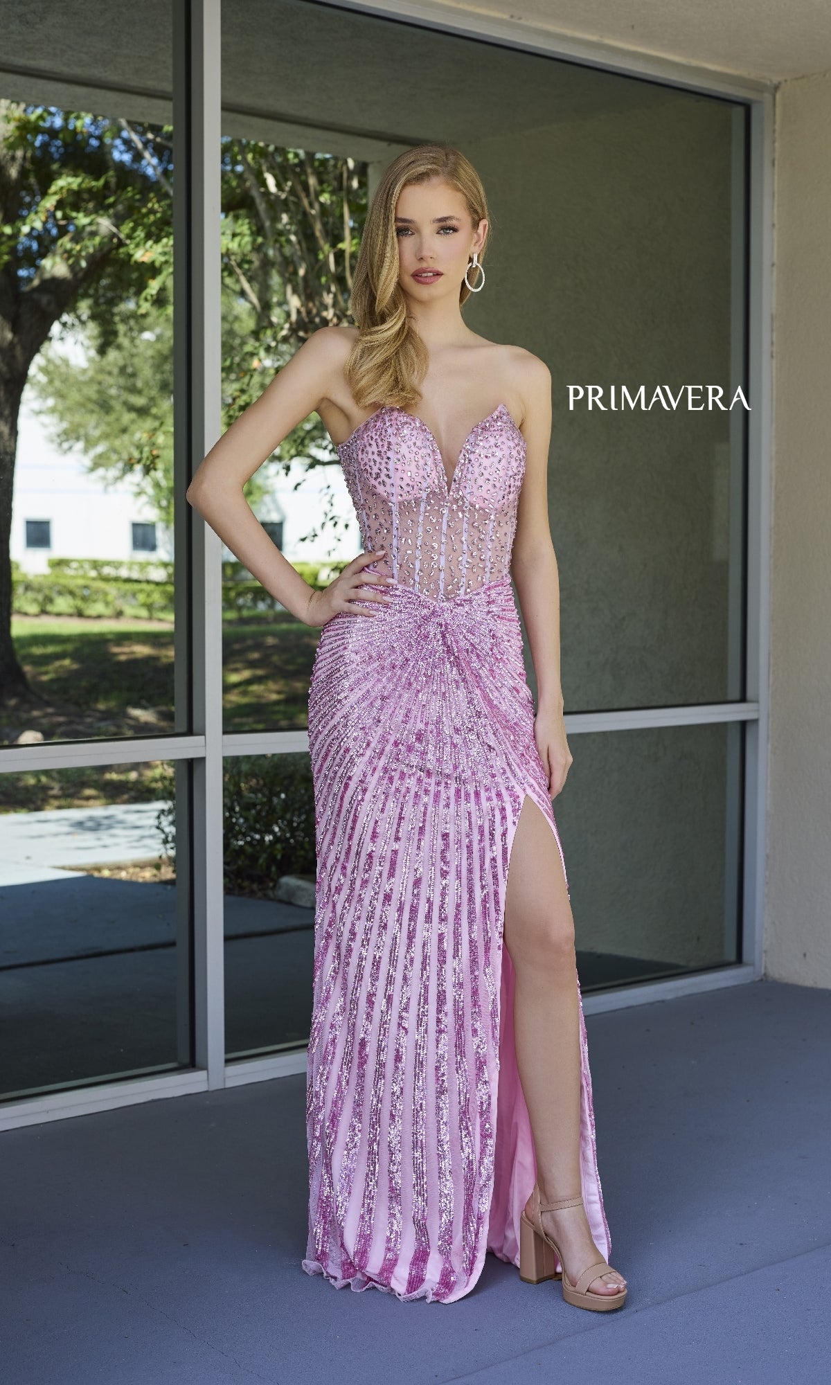 A woman stands outside by glass windows wearing the Strapless Long Sequin Prom Dress: Primavera 4304 in pink and silver with a thigh-high slit.