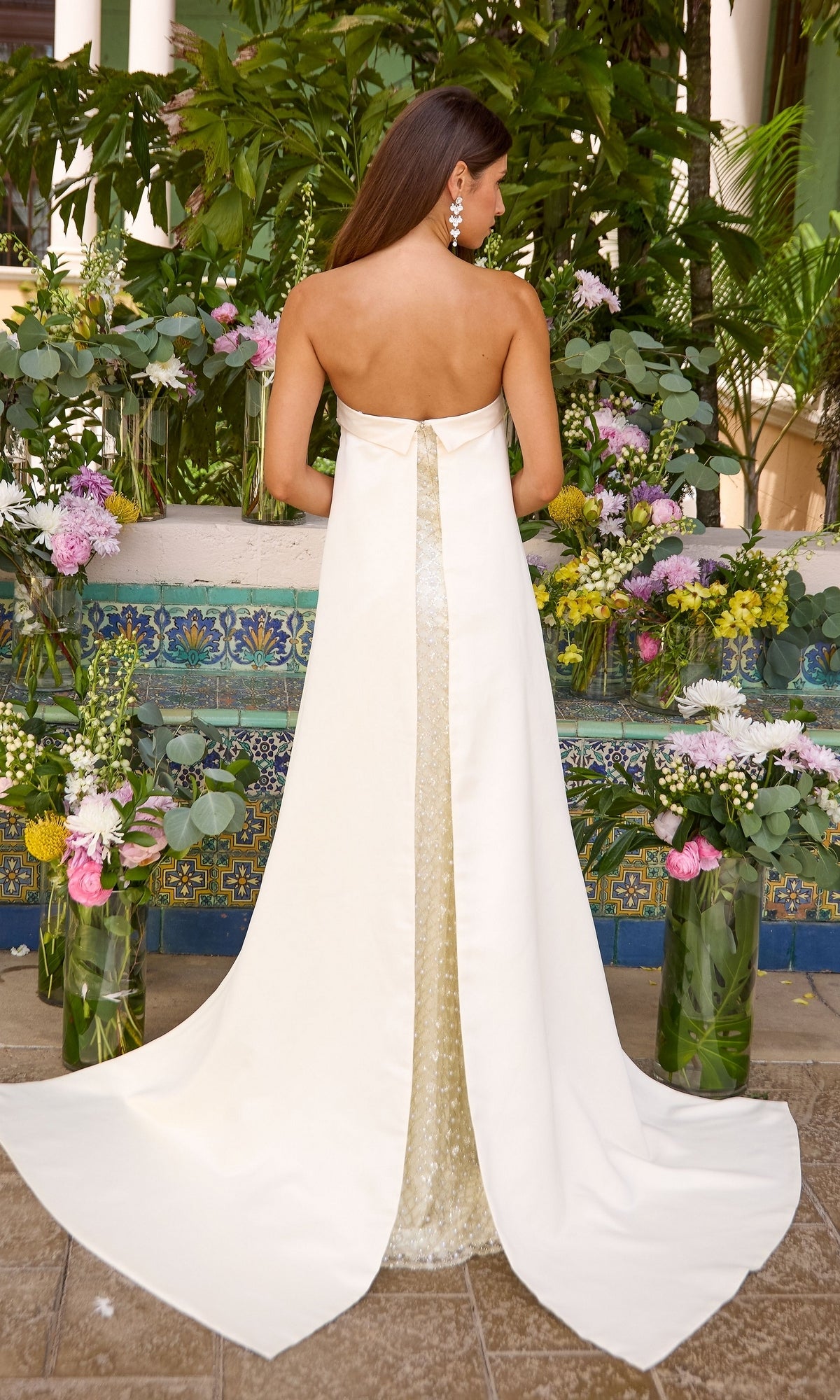 Back view of a white, floor-length Ava Presley 43060 strapless dress with a sequined center panel and small train, standing in front of floral arrangements.