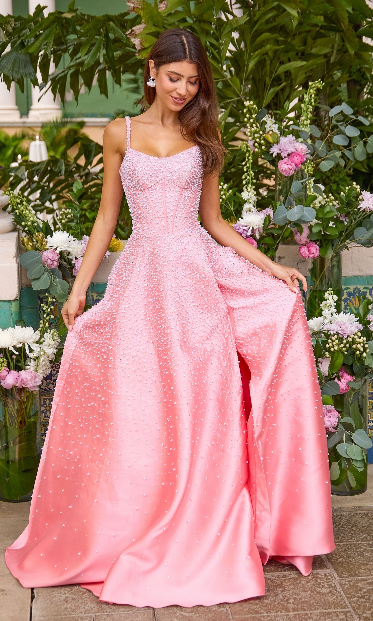 A woman wearing the Long Prom Dress: Ava Presley 43065, a pink pearl gown with a corset-style bodice and scattered embellishments, standing in front of a floral arrangement.