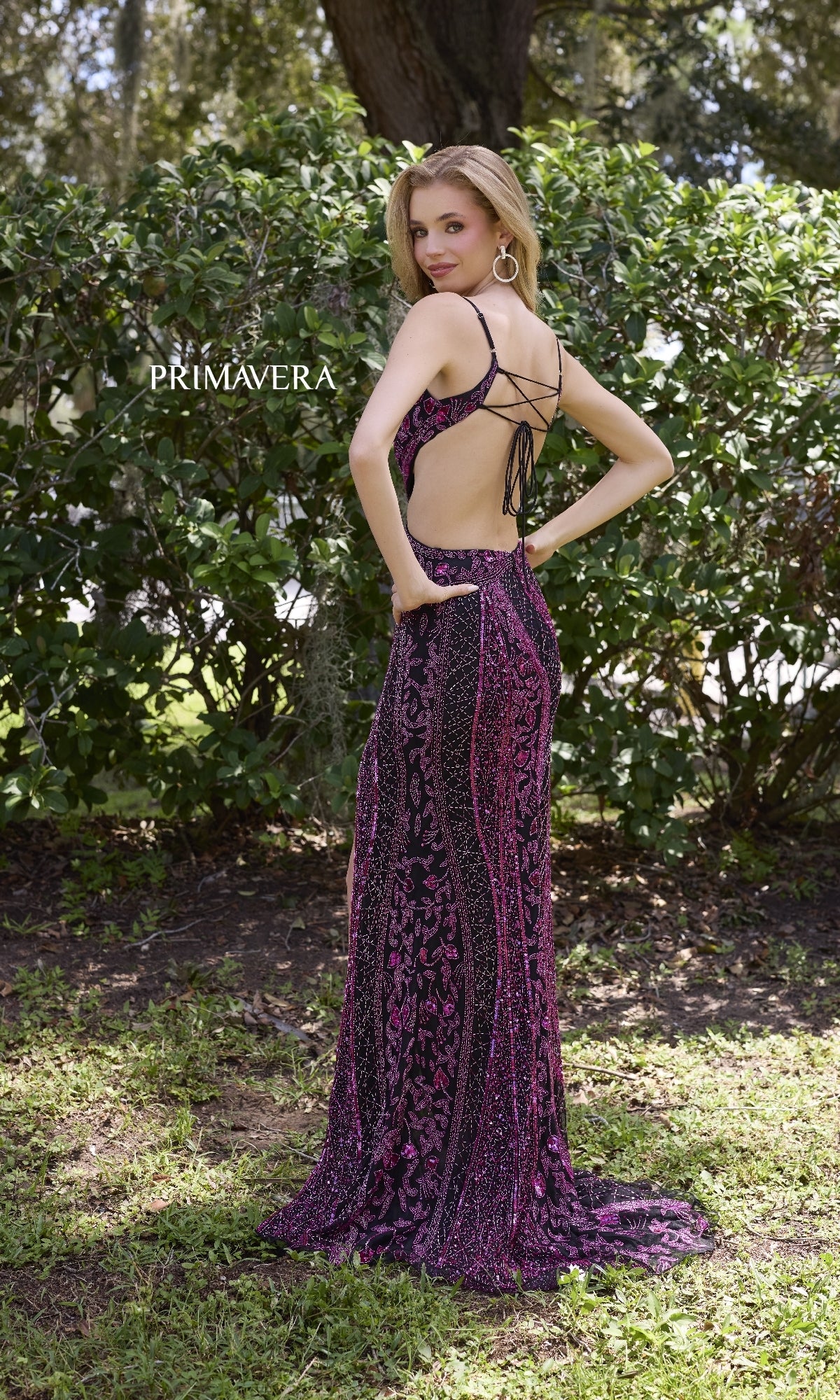 A woman stands outdoors in the Primavera 4308 Long Prom Dress, a floor-length gown with purple and black patterns, thin straps, and an open back. Shrubs and trees are behind her.