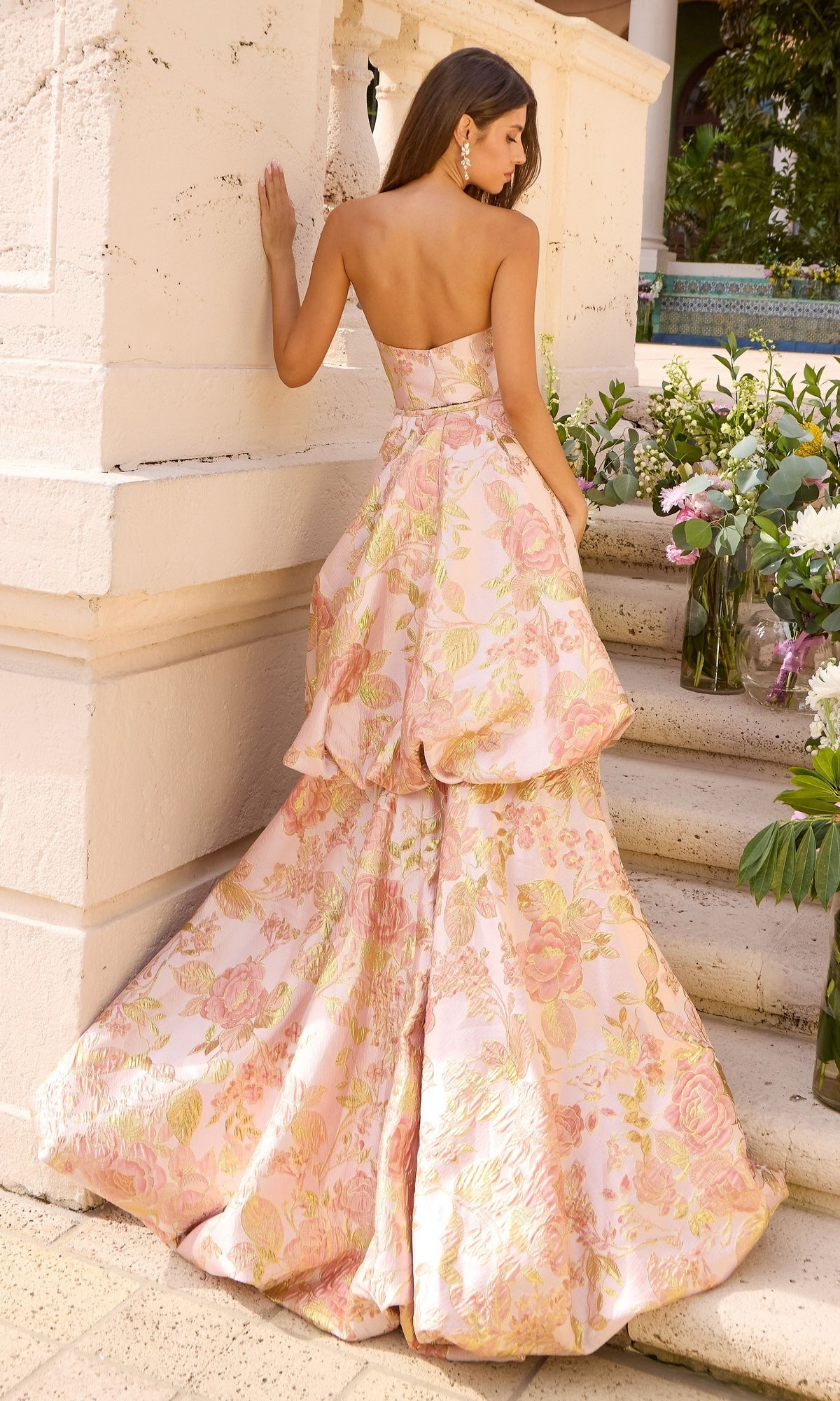 A woman wearing a Long Prom Dress: Ava Presley 43091, a strapless, floor-length metallic mermaid gown with a pink and gold floral brocade pattern, standing on stone steps.