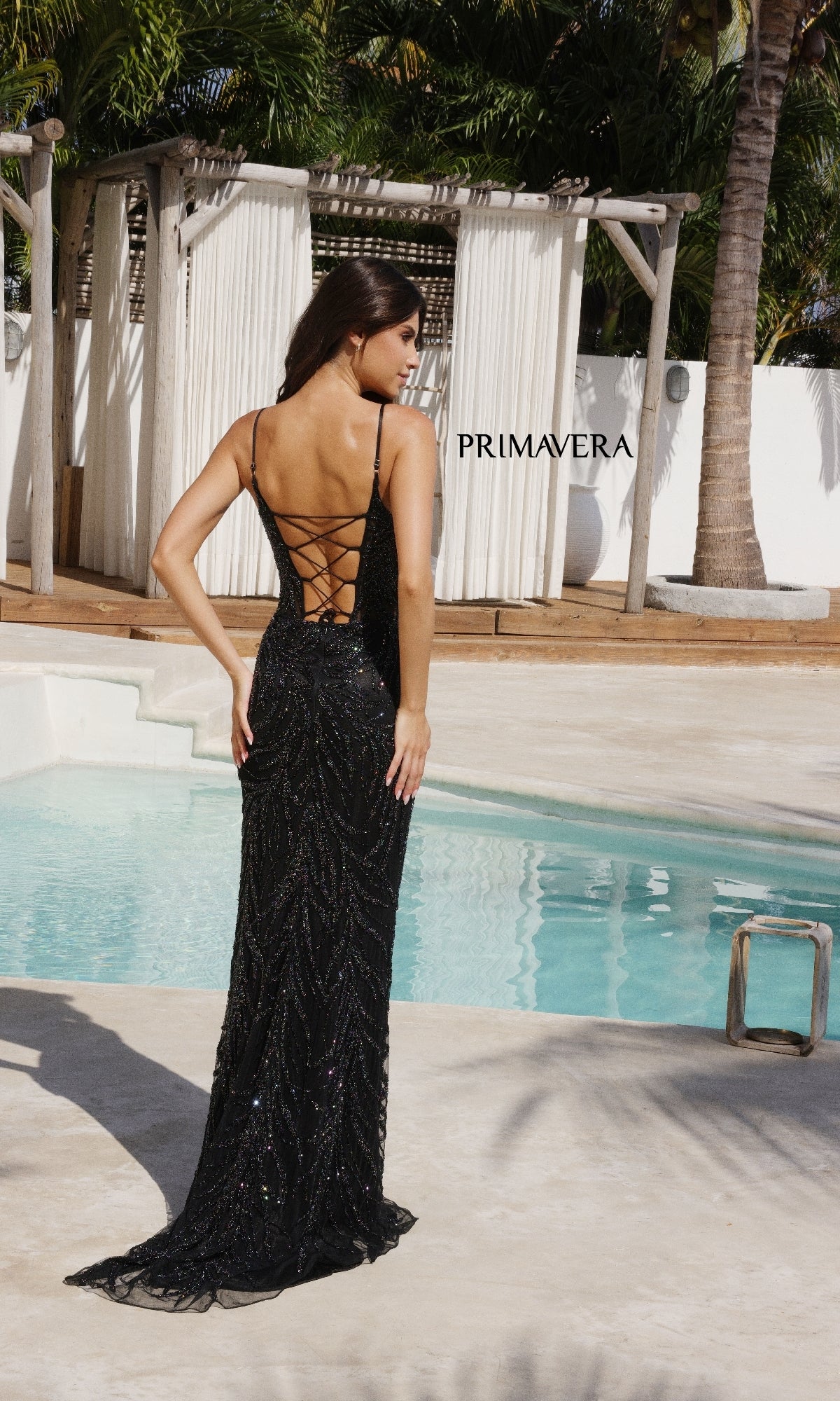A woman in the Primavera 4310 Scoop-Neck Long Beaded Prom Dress stands by a pool, facing away, with lush greenery and white outdoor curtains forming an elegant backdrop.